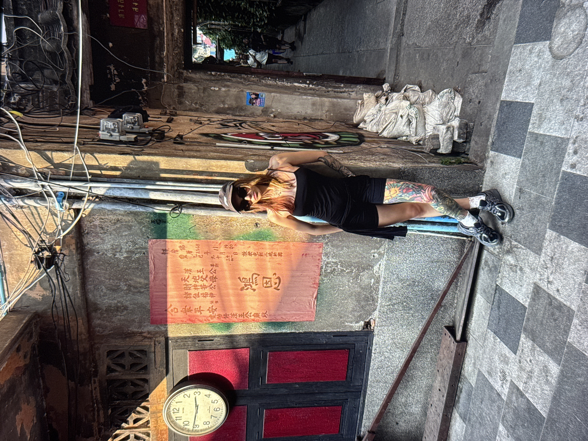 A woman with tattoos on her legs and arms stands leaning against a weathered concrete wall. She is wearing a black tank top, black shorts, a baseball cap, sunglasses, and black sneakers. The wall has a red poster with Chinese characters, electrical wires, and a clock mounted on it. The ground is paved with gray and black square tiles. To the right, a narrow alleyway with buildings and people can be seen. The scene appears to be in an urban environment, possibly in Asia, given the script on the poster.