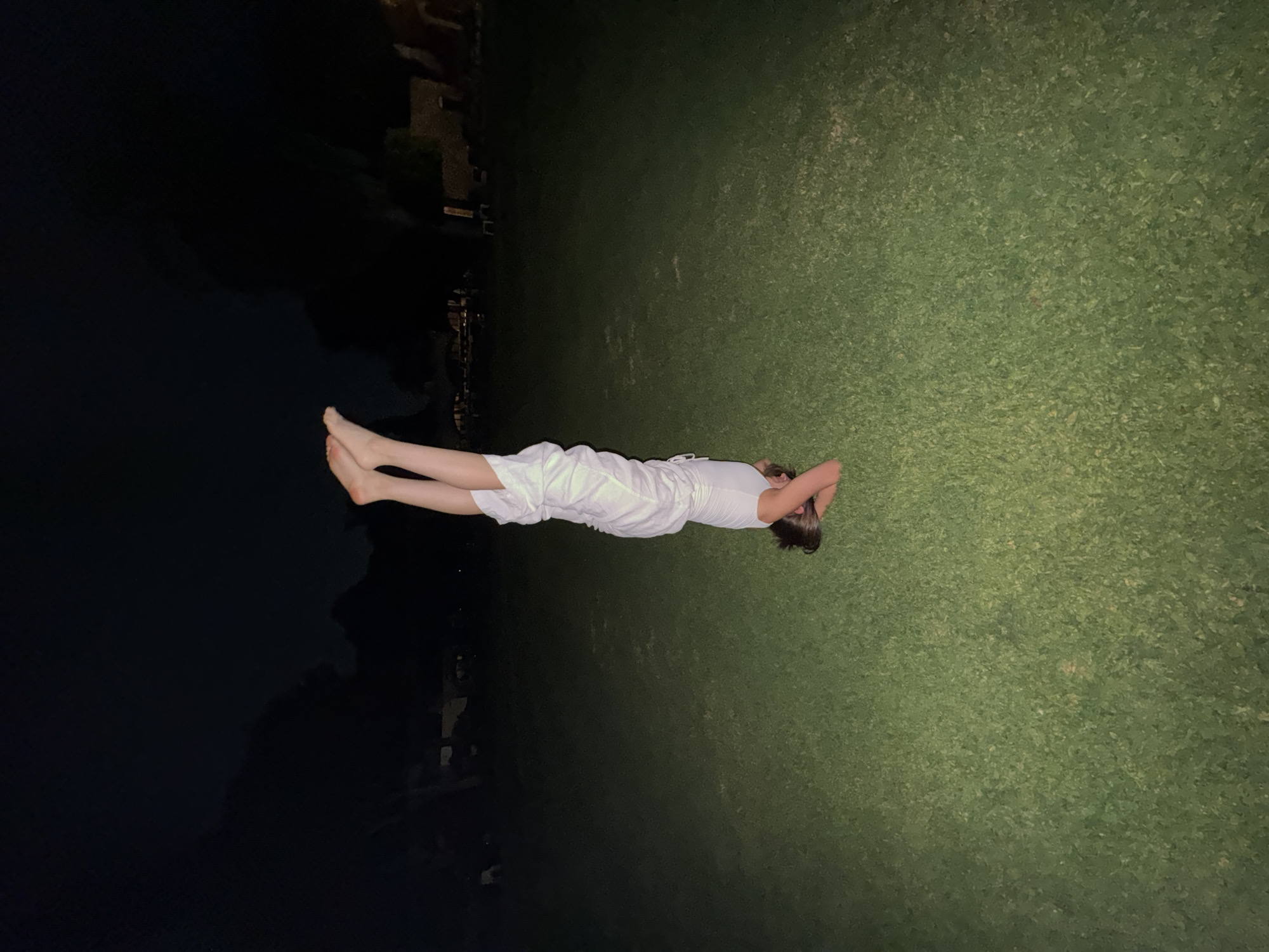 A person is lying down on grass next to a dark body of water or shadowed area. The person is wearing white clothing. The environment appears to be dimly lit, with possible building structures visible in the background. This image may be set in Cocoyoc, Mexico.