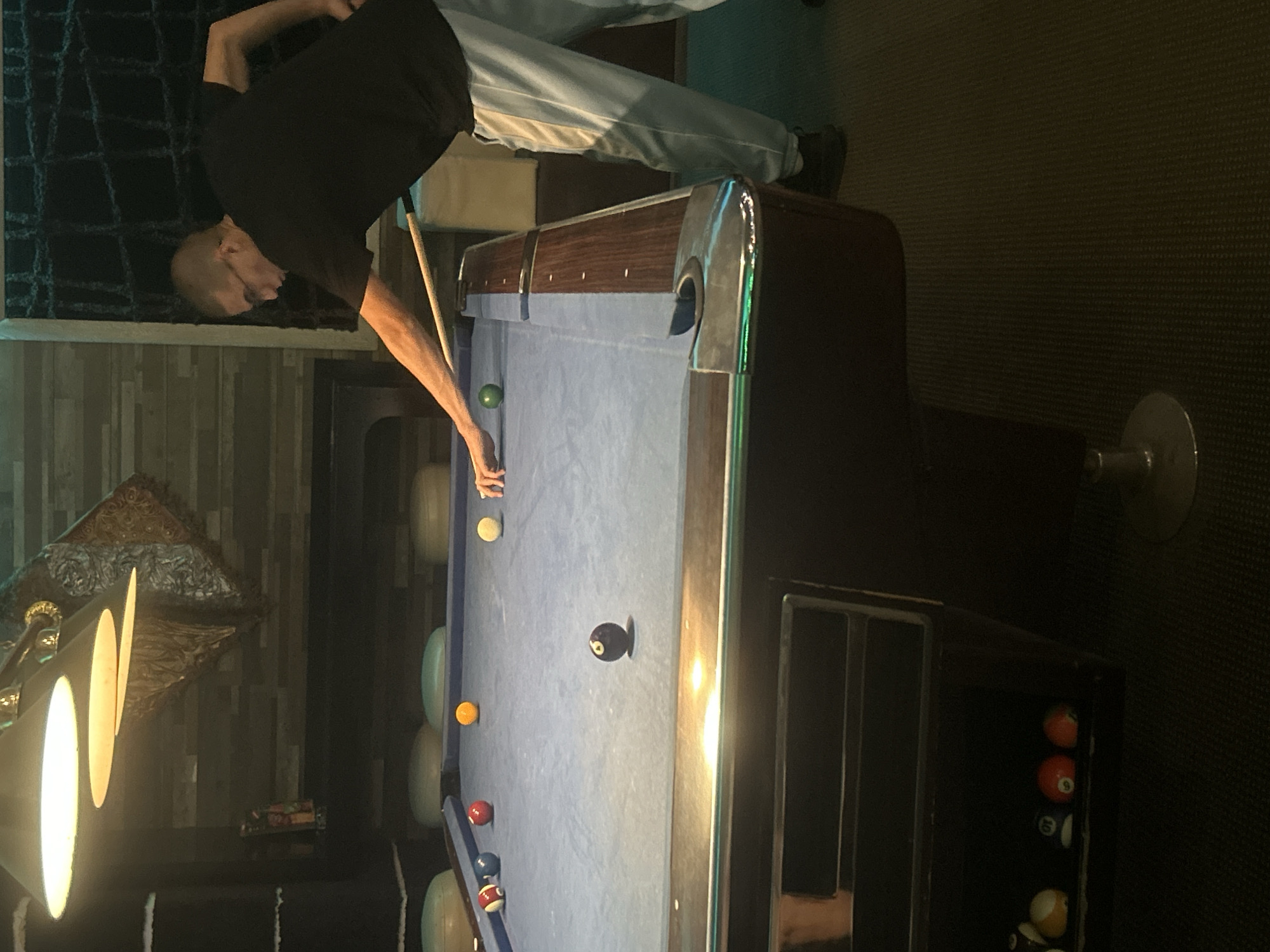 A man is playing billiards. He is bald and wearing a black t-shirt and light grey pants. The billiard table is light blue with dark brown trim and silver corner pieces. The table has several billiard balls on it. The environment includes a chandelier, dark flooring, and a storage cabinet with billiard balls inside. There is no indication of the location being Pleven, Bulgaria.