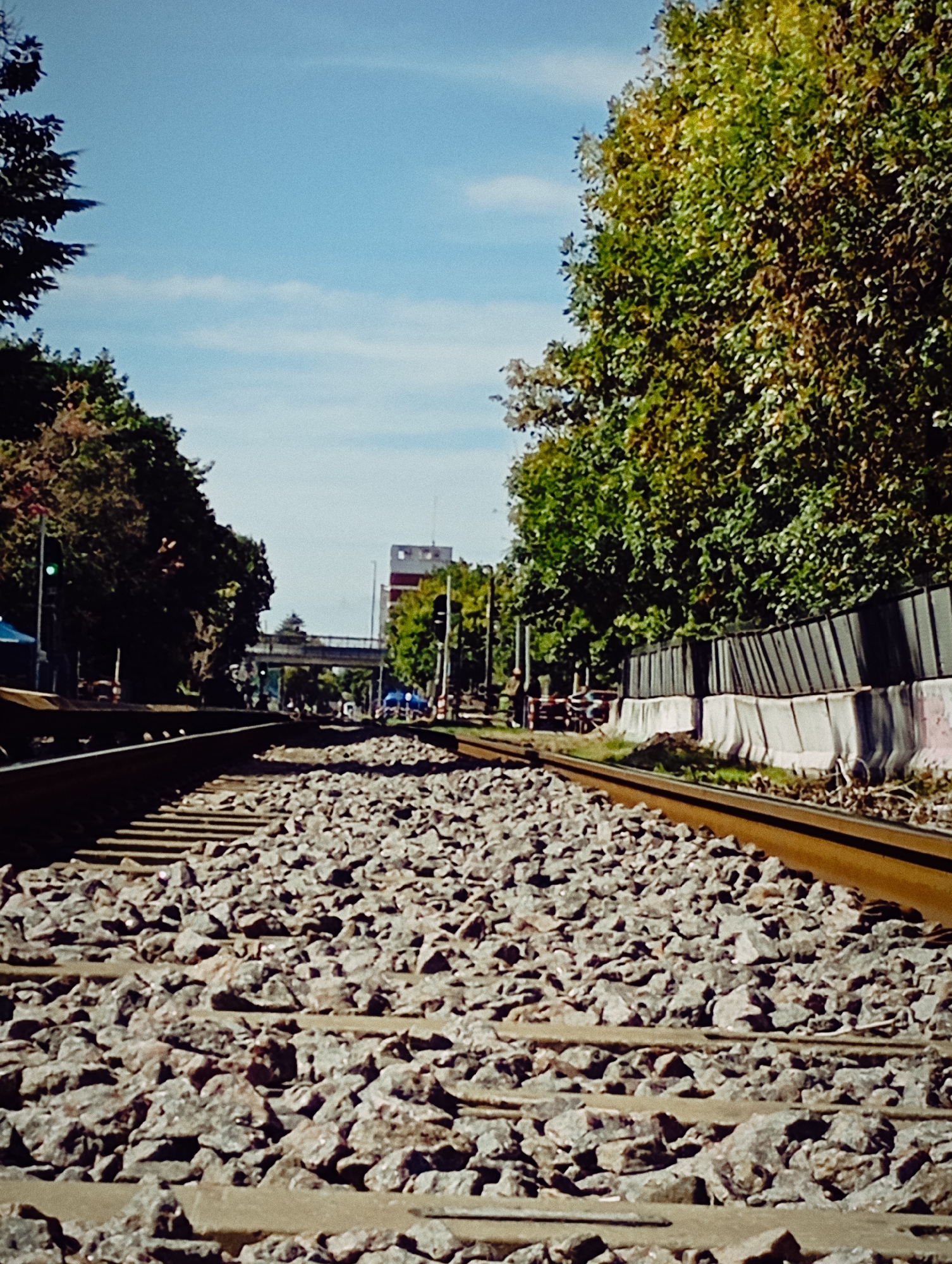 The image depicts a section of railway tracks in Buenos Aires, Argentina. The tracks are laid on a bed of gravel. Vegetation borders either side, with a fence on the right. In the background, a building and an overpass are visible. There are trees on either side of the tracks. Traffic lights are visible, as are cars on the street. The sky is blue with some clouds.