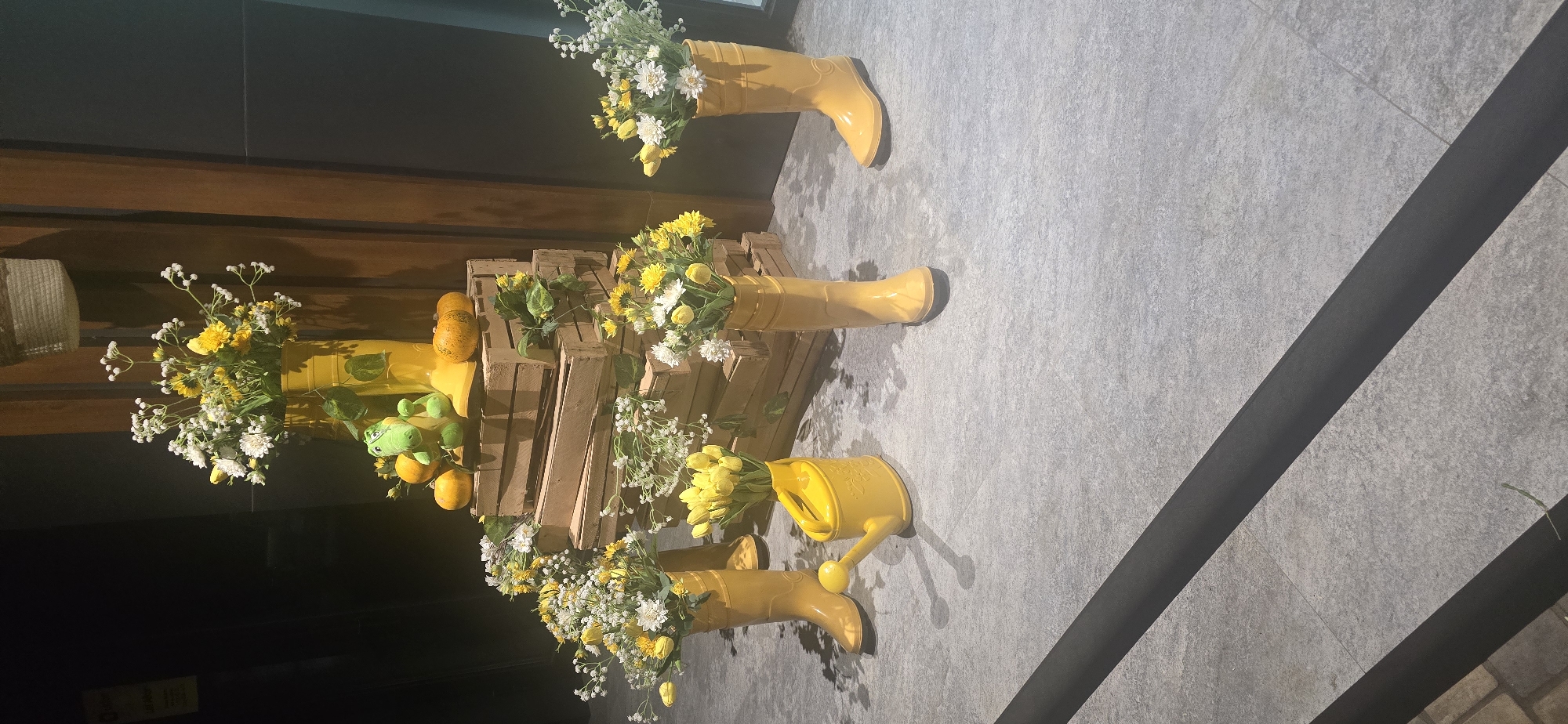 This image shows an outdoor decorative display in Tashkent, Uzbekistan, likely for a seasonal event. The primary elements are several yellow rubber boots used as planters for bouquets of yellow and white flowers. These boots are arranged around a tiered structure made of wooden crates. Among the flowers and crates, there are small pumpkins and a stuffed green alligator toy. The ground is paved with gray tiles, and a dark, raised barrier runs diagonally across the foreground. The background is dark and indistinct, suggesting an indoor or shaded area. The overall impression is a whimsical, summery or autumnal display.