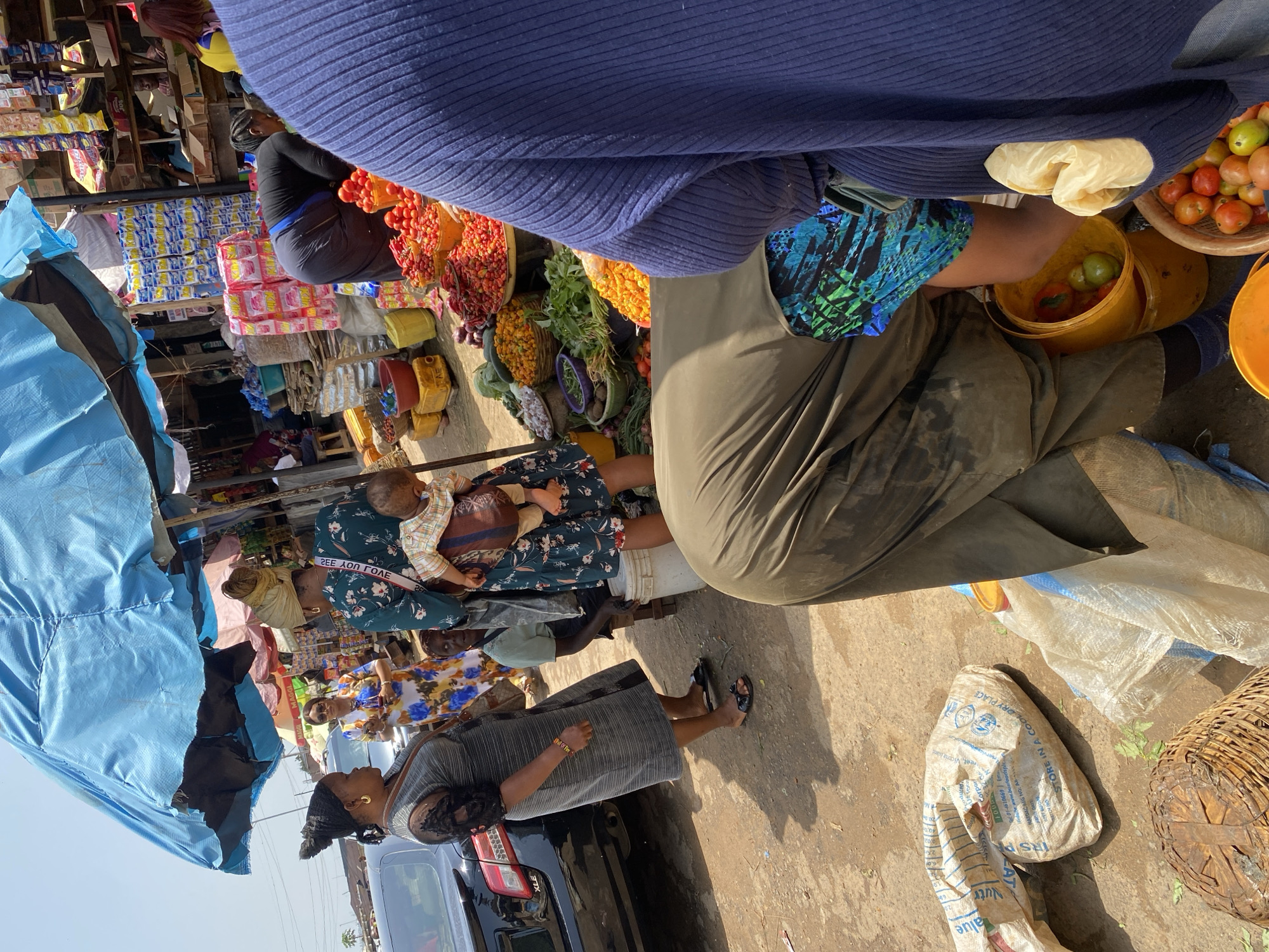 This image depicts a bustling market scene in Jos, Nigeria, under a large blue tarp providing shade. In the foreground, a person wearing a dark blue ribbed sweater is partially visible on the right. In the center, another individual, clad in olive green trousers and a patterned blue and green skirt, is bent over a yellow bucket containing tomatoes and peppers. To their left, a wooden basket is filled with tomatoes, and another yellow bucket is visible. In the background, various market stalls display an abundance of fresh produce, including tomatoes, peppers, cucumbers, and leafy greens. Several individuals are engaged in shopping and selling activities. A woman in a grey dress stands near a black car, while another woman in a patterned dress is seen with a baby strapped to her back. The ground is dusty, typical of an outdoor market.