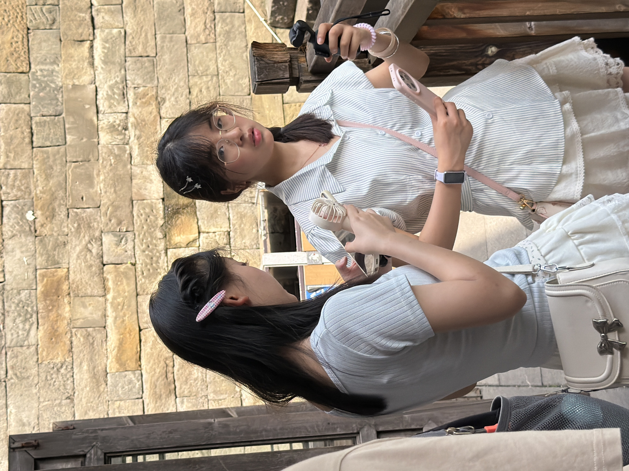 In Yuzhong District, China, two young East Asian women stand against a weathered brick wall. The woman on the left, with long black hair and a light blue ribbed t-shirt, holds a smartphone and a white handbag with a bow clasp. The woman on the right, wearing a striped shirt and glasses, holds a black gimbal and a pink smartphone, with a pink cross-body bag. A small white fan is also visible.