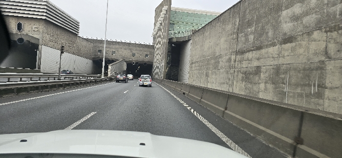 The image presents a view from the driver's perspective on a multi-lane highway or major road, indicating ongoing travel. In the foreground, the white hood and a portion of the dashboard of the vehicle are visible. Several cars are seen driving ahead on the asphalt road, which features clear white lane markings and is bordered by concrete barriers and guardrails.
To the left, the road runs alongside a complex of imposing concrete structures. A large building with distinctive horizontal louvers on its upper section dominates the immediate left, with a wide tunnel entrance directly beneath it. Further down this side, another tunnel entrance and more grey concrete structures are visible. A traffic light stands next to the first tunnel entry.
On the right side of the road, a tall, unadorned concrete wall stretches into the distance. Beyond this, a large, modern concrete structure with a patterned, possibly perforated or glass-covered green facade becomes prominent, appearing to integrate with another large opening or a downward-sloping ramp.
The sky is overcast, suggesting a cloudy day, and the lighting is flat. The overall setting is characterized by massive, functional infrastructure, typical of an industrial or transportation hub like Vondelingenplaat in the Netherlands, where this image was captured. No discernible text is visible in the frame.