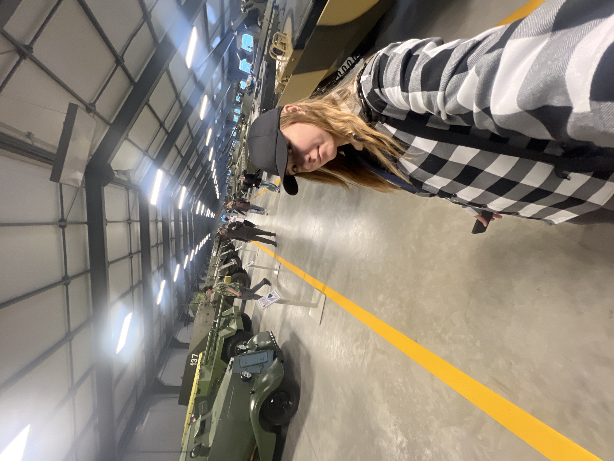 This media file shows a young woman with blonde hair, wearing a black baseball cap and a black and white buffalo plaid shirt, taking a selfie. She is in a large indoor space that appears to be a museum or exhibition hall, possibly for military vehicles. In the background, several green military vehicles are displayed, lined up along a concrete floor marked with a yellow line. Other visitors can be seen observing the vehicles. The lighting suggests it is daytime, with artificial lights from the ceiling illuminating the space. The woman's expression appears casual and engaged with the camera. The vehicles are arranged in a row, and some have numbers or markings on them, such as "137" visible on one. The overall setting is a well-lit, spacious indoor area designed for public viewing of exhibits. The location is identified as Kubinka, Russia.