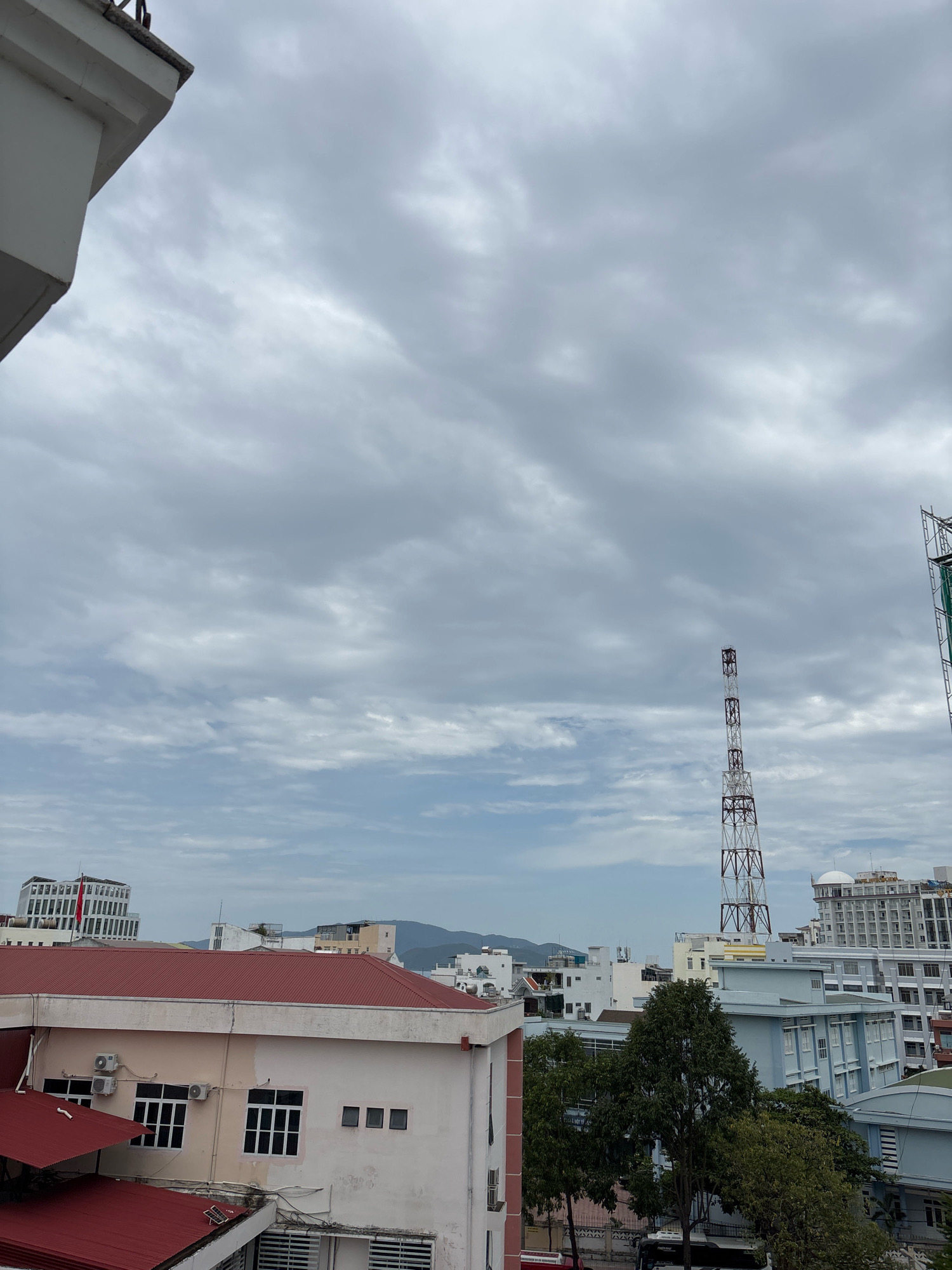 The image is a view of a city skyline with a cloudy sky. In the foreground, there is a section of a white building with a concrete railing. The middle ground of the image features a red-roofed building with multiple windows, some of which are air-conditioned units. To the right of this building, there are several other buildings of varying heights and architectural styles, including a tall, lattice-style radio tower with red and white banding. Trees are visible between some of the buildings. In the background, more buildings and distant hills or mountains can be seen. The overall impression is an urban environment under an overcast sky.