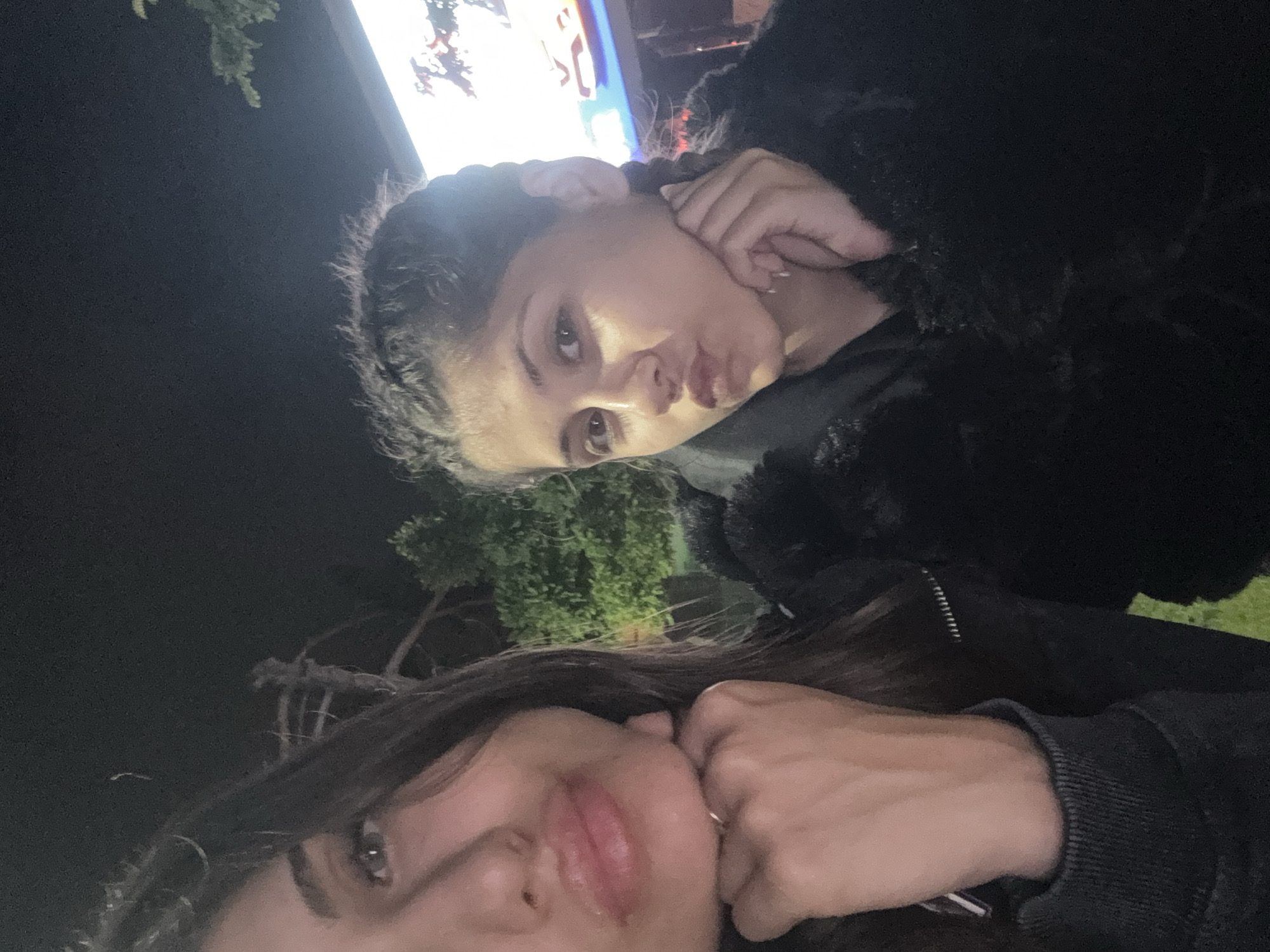 The image displays two females positioned closely together during nighttime in Nesebar, Bulgaria. The female on the left, partially cropped, has long dark hair and light-colored eyes. She wears a dark long-sleeved top with ribbed cuffs, and her left hand, adorned with a ring on the ring finger, rests beneath her chin. Her lips are notably pursed. The female on the right is fully visible, with dark hair styled in two braids and light-colored eyes. She wears a dark, possibly textured or faux-fur, jacket. Her right hand is also placed beneath her chin, and her lips are pursed, mirroring the pose of the other individual. The background is dark, indicating night, with visible foliage and trees. On the far right, a brightly illuminated outdoor sign or screen, displaying indistinct red and blue elements, is visible, suggesting a commercial area.