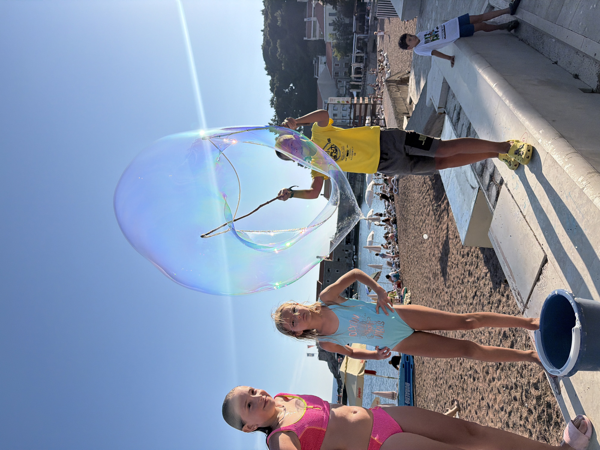 In Petrovac centar, Montenegro, on a sunny day at the beach, four children are engaged in making a large bubble. A boy in a yellow t-shirt and grey shorts is holding a stick with a loop, immersed in a bucket of bubble solution, and is in the process of creating a massive, iridescent bubble. To his left, two girls in swimsuits stand on the sand. The girl on the left wears a pink and yellow bikini, and the girl on the right wears a light blue swimsuit. A younger boy in a white t-shirt and blue shorts stands on a raised concrete structure to the right, looking towards the bubble. The beach is populated with beachgoers lounging on chairs under umbrellas, and the Adriatic Sea is visible in the background, with buildings and a fortified structure on a hill overlooking the scene. The sky is clear and blue.