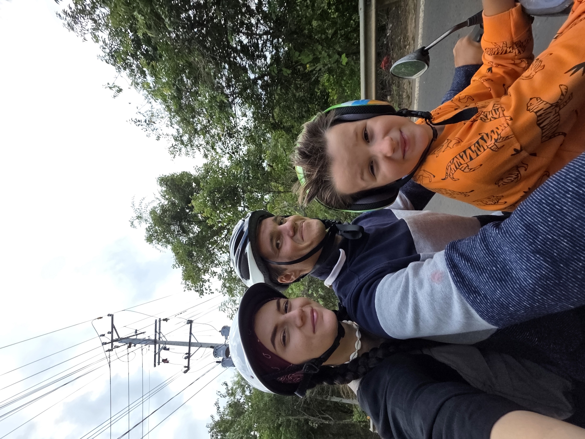 Three people are on a motor scooter in Thôn 5, Vietnam. The front person is wearing a white helmet, a black shirt, and a shell necklace. The middle person is wearing a white helmet and a blue and white shirt. The person in the back is wearing a helmet and an orange shirt with tigers on it. The scooter has a mirror. Powerlines and trees are visible in the background. The sky is overcast.