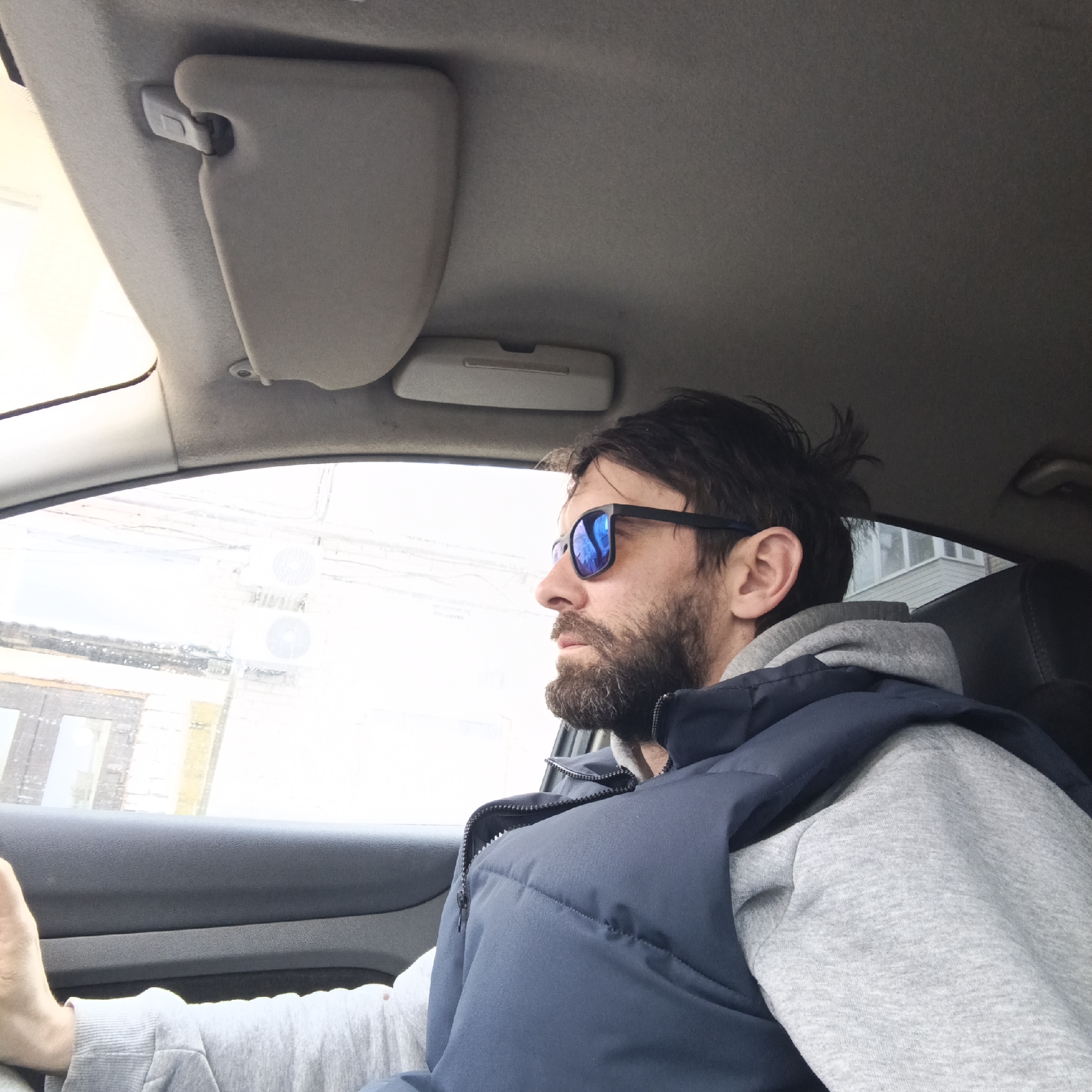 A medium shot depicts a man in the driver's seat of a car. He is wearing blue sunglasses, a grey hooded sweatshirt, and a navy blue vest. He has dark hair and a beard. The sun visor is down on the driver's side. Outside the car window, blurred buildings suggest a city location. Based on visual cues like the architecture and potential Cyrillic characters visible through the window, the location could be Chaikovsky, Russia. The man is facing forward, seemingly concentrating on the road. His hand is slightly visible, suggesting he is holding the steering wheel.