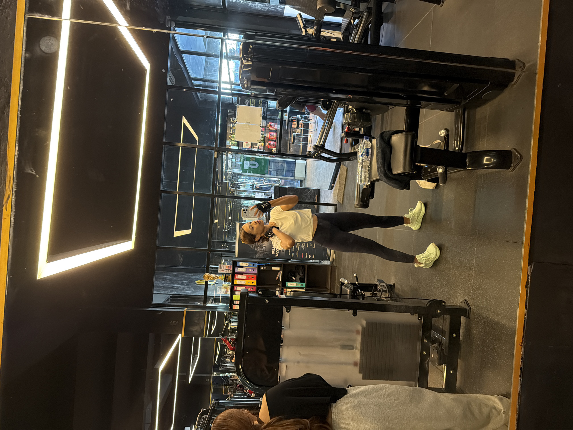 The image is a distorted, slightly off-center view of a gym, likely taken through a mirror. The reflection shows two individuals, a woman exercising and another person sitting down. The exercising woman is dressed in a white top and black leggings, and she appears to be using her phone while working out. Her sneakers are light green. Gym equipment, including a seated leg press machine and cable machines, is visible in the foreground and background. The interior features black and glass walls, with fluorescent lighting in geometric patterns. Outside the glass walls is a view of Denpasar, Indonesia, where signs and storefronts are visible, suggesting a location in an urban area.