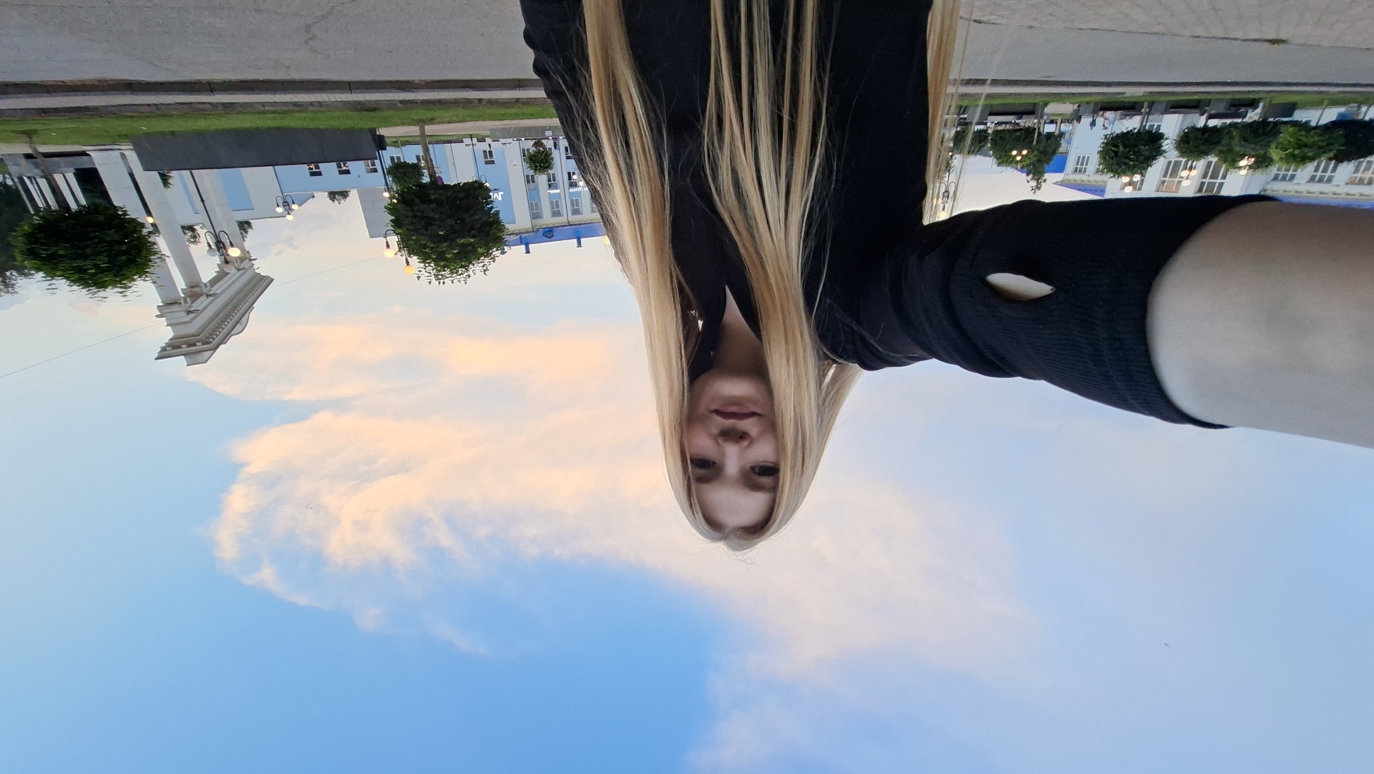 A young woman with long blonde hair is taking a selfie outdoors in Chaikovsky, Russia. She is wearing a black long-sleeved top with a cut-out detail on the left sleeve. The background features a bright blue sky with wispy pink and orange clouds, suggesting it is either sunrise or sunset. To the right of the woman, there is a large white architectural structure with columns and an archway, possibly a monument or part of a building. In the distance, a light blue building with white trim is visible, along with several trees and streetlights. A paved road and sidewalk are in the foreground.