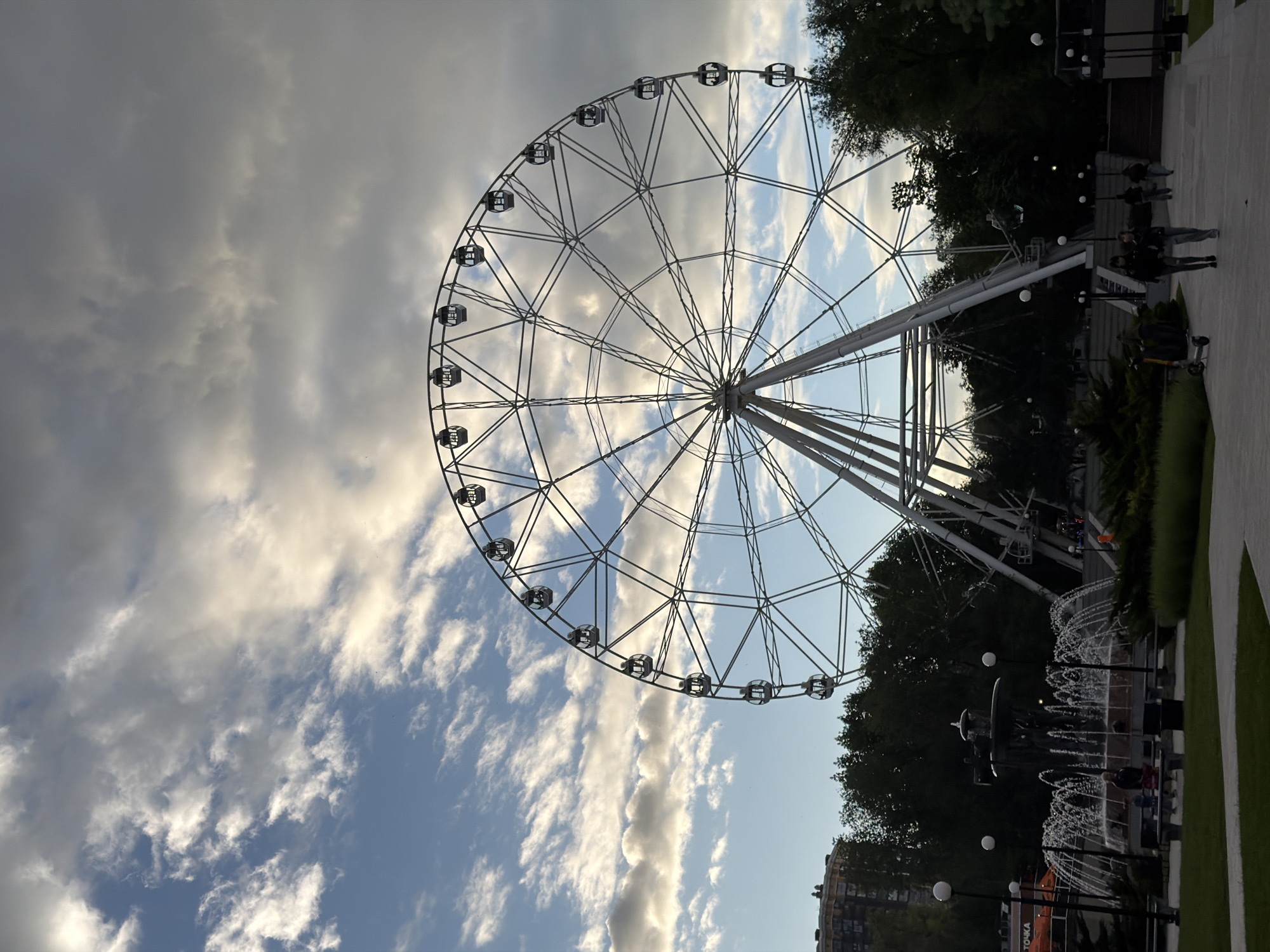 A ground-level, rotated shot depicts a large Ferris wheel in Rostov-on-Don, Russia, set against a backdrop of a cloudy sky. The Ferris wheel is primarily white with a metal framework forming the wheel structure and individual passenger cabins. The sky shows varying cloud formations from light, scattered clouds to denser, gray cloud cover. In the lower right, there are trees, bushes, and a pedestrian walkway. Fountains are visible near the base of the Ferris wheel. People are walking along the path. The overall lighting suggests it is either early morning or late afternoon.