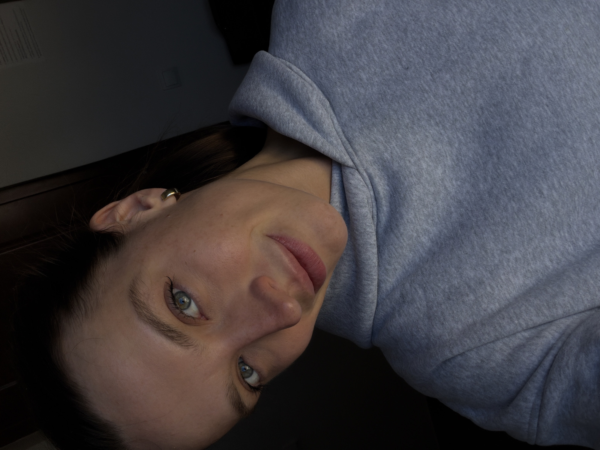 A woman with fair skin, light blue eyes, and dark hair pulled back into a bun, is wearing a gray hoodie. The photo appears to be taken indoors, with a dark wall visible in the background. The image is rotated, with the woman's face angled to the lower left. A gold earring is visible in her left ear. No specific location or city is discernible from the image.