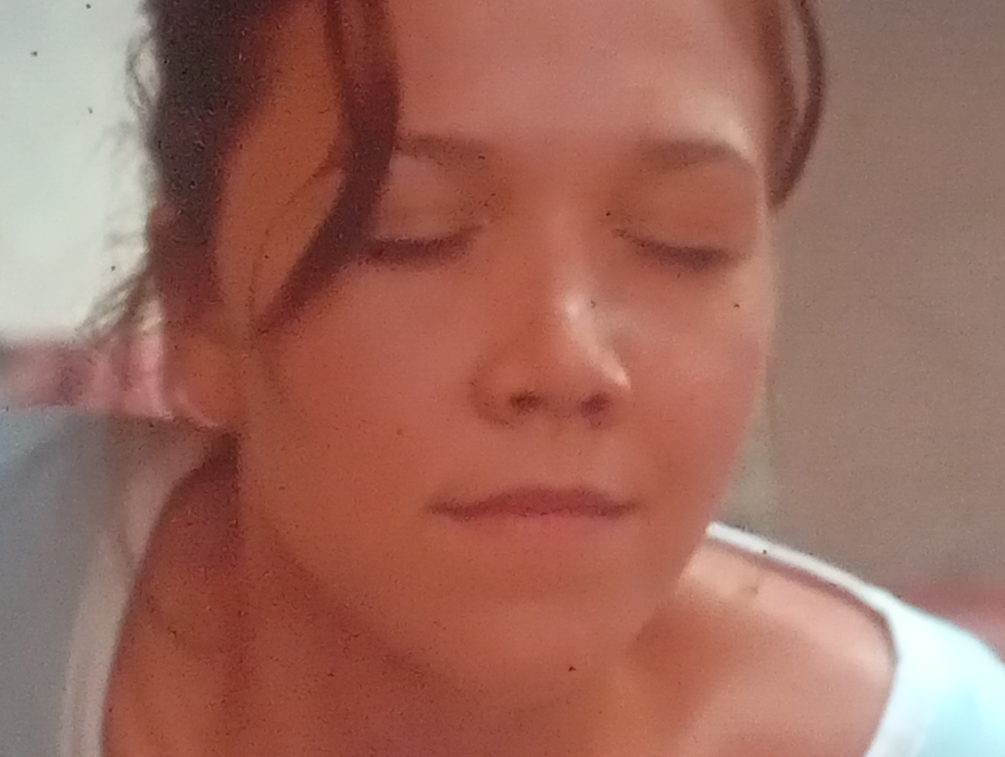 This is a close-up, slightly blurry, over-the-shoulder shot of a young woman with her eyes closed. She has light brown hair that is pulled back. Her face is lit from the front. The background is a neutral, light brown color. The image was captured in Sofiivska Borshchahivka, Ukraine.