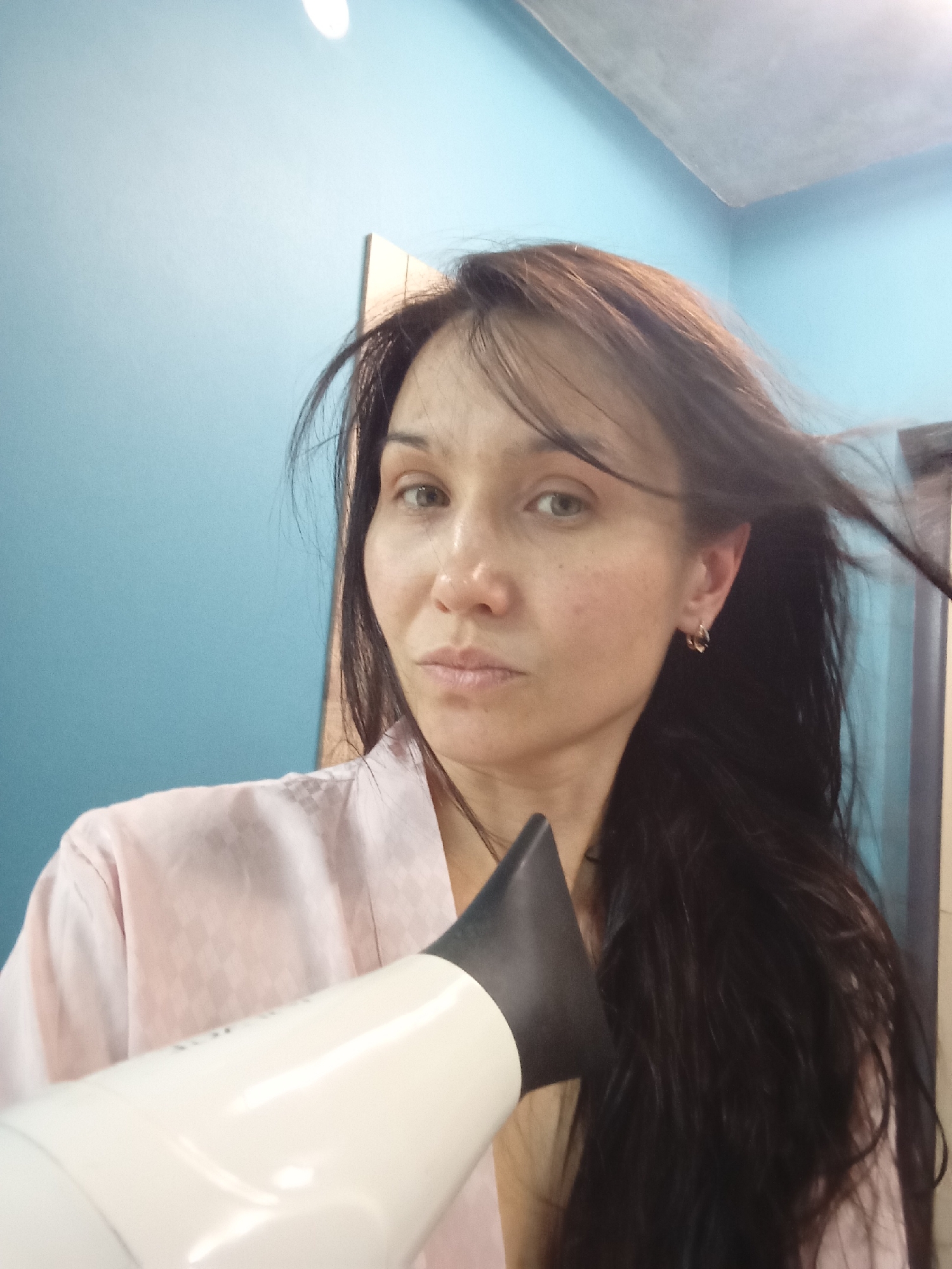 A woman with long dark hair is holding a white hair dryer with a black nozzle close to her face, presumably drying her hair. She is wearing a pink robe. The background consists of a light blue wall. This photo appears to be taken indoors. The location is specified as Pervomaysky, Russia, but no details in the image confirm this.