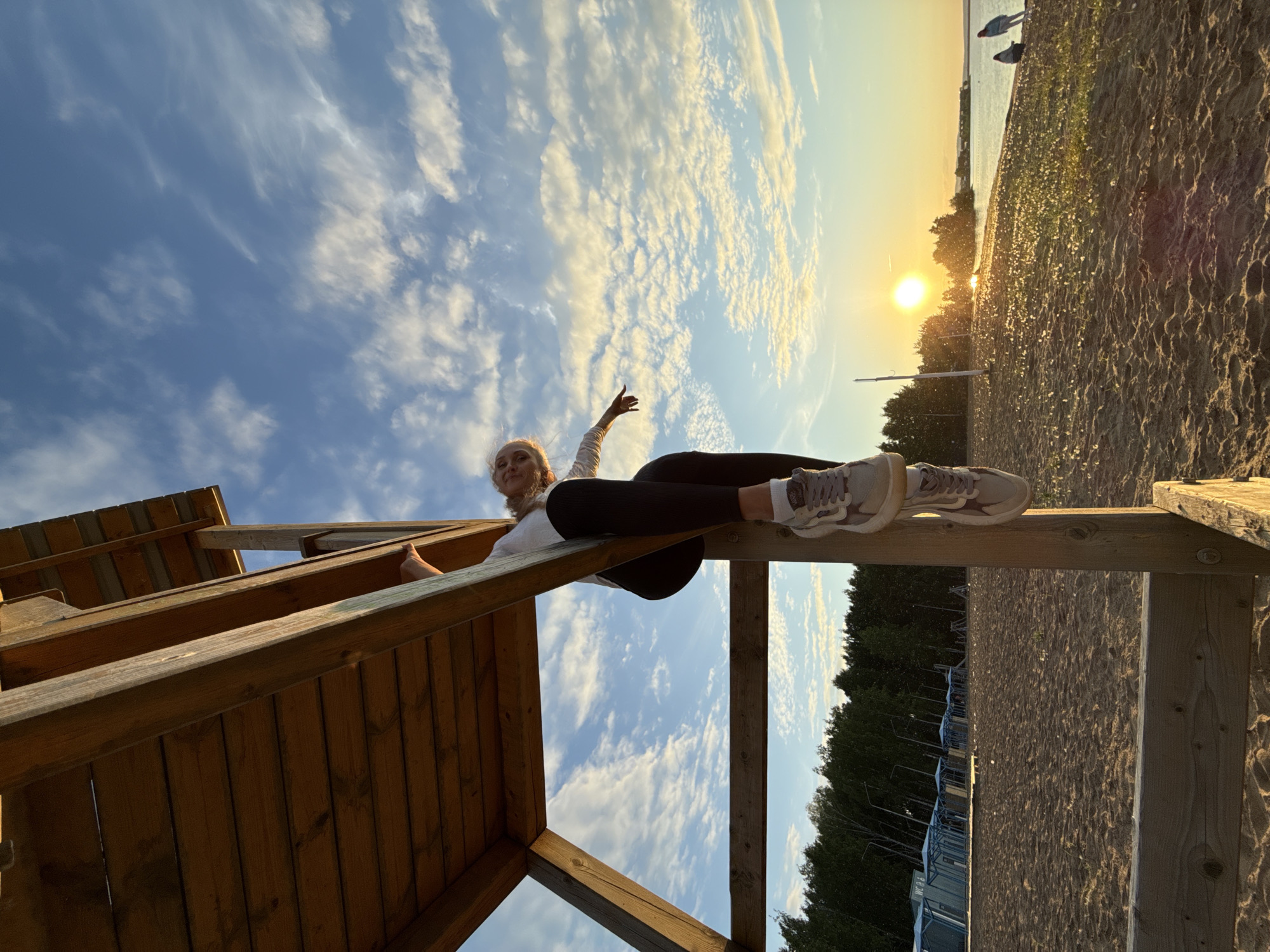 A low-angle outdoor photograph captures a person hanging from a wooden structure on a sandy beach during sunset, likely on the Votkinsk Reservoir in Chaikovsky, Russia.
The central subject, a person with light-colored hair, is positioned on the right side of the frame, suspended from a horizontal beam of a robust wooden structure. They are wearing a light-colored long-sleeved shirt, dark leggings, and light-toned sneakers with white socks. Their left arm is extended, index finger pointing towards the horizon, while their right arm supports their weight. Their legs are draped over a vertical support beam.
The wooden structure, constructed from dark, weathered planks and beams, occupies the left and central-right portions of the frame. Its underside and structural elements are prominent, suggesting it is a multi-tiered platform, possibly a lifeguard tower or observation point.
The environment depicts a sandy beach stretching from the foreground to a large body of water on the right. The sun, low on the horizon, casts a bright, warm yellow glow across the water and into the sky, indicating a sunset. Two distant figures are visible near the water's edge on the far right. The sand exhibits textural variations and subtle ripples. On the left side of the beach, a row of blue changing cabins or small kiosks is visible, alongside folded beach umbrellas. A dense line of deciduous trees forms the treeline behind these amenities.
The sky above is predominantly blue, transitioning to shades of orange and yellow near the horizon where the sun is setting. Scattered white and light grey clouds are present throughout the sky, illuminated by the sunset, with denser cloud formations near the horizon.