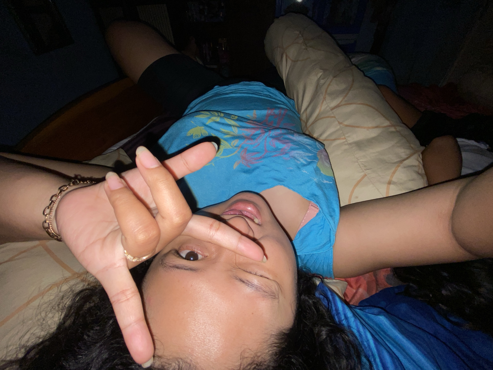 This is a first-person, upside-down selfie of a young woman lying on a bed in what appears to be a bedroom. She is wearing a blue t-shirt with a floral print and dark shorts. Her left hand is raised to her face, with her index and middle fingers forming a peace sign, and her right hand is resting on her thigh. She has dark curly hair and is wearing a gold bracelet and a ring. Her expression is playful, with one eye winking and her mouth slightly open. The background is dimly lit, suggesting it is nighttime or the room is dark. The bed is covered with pillows and blankets, one of which has a patterned design. There is no visible text in the image. The context provided suggests the location is Gelam, Indonesia.