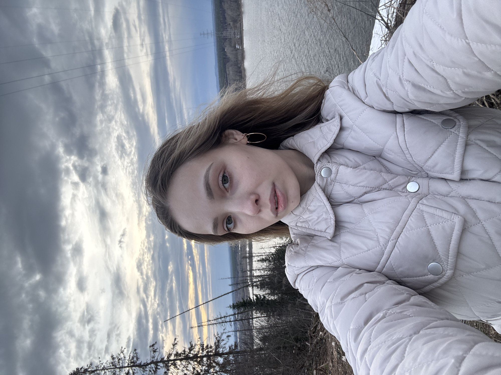 A young woman is taking a selfie outdoors in Chaykovsky, Russia. She wears a light-colored quilted jacket and gold hoop earrings. Behind her, the scene features a body of water reflecting a cloudy sky. Power lines are visible in the distance, along with trees and vegetation along the shoreline. The foreground includes dry vegetation.