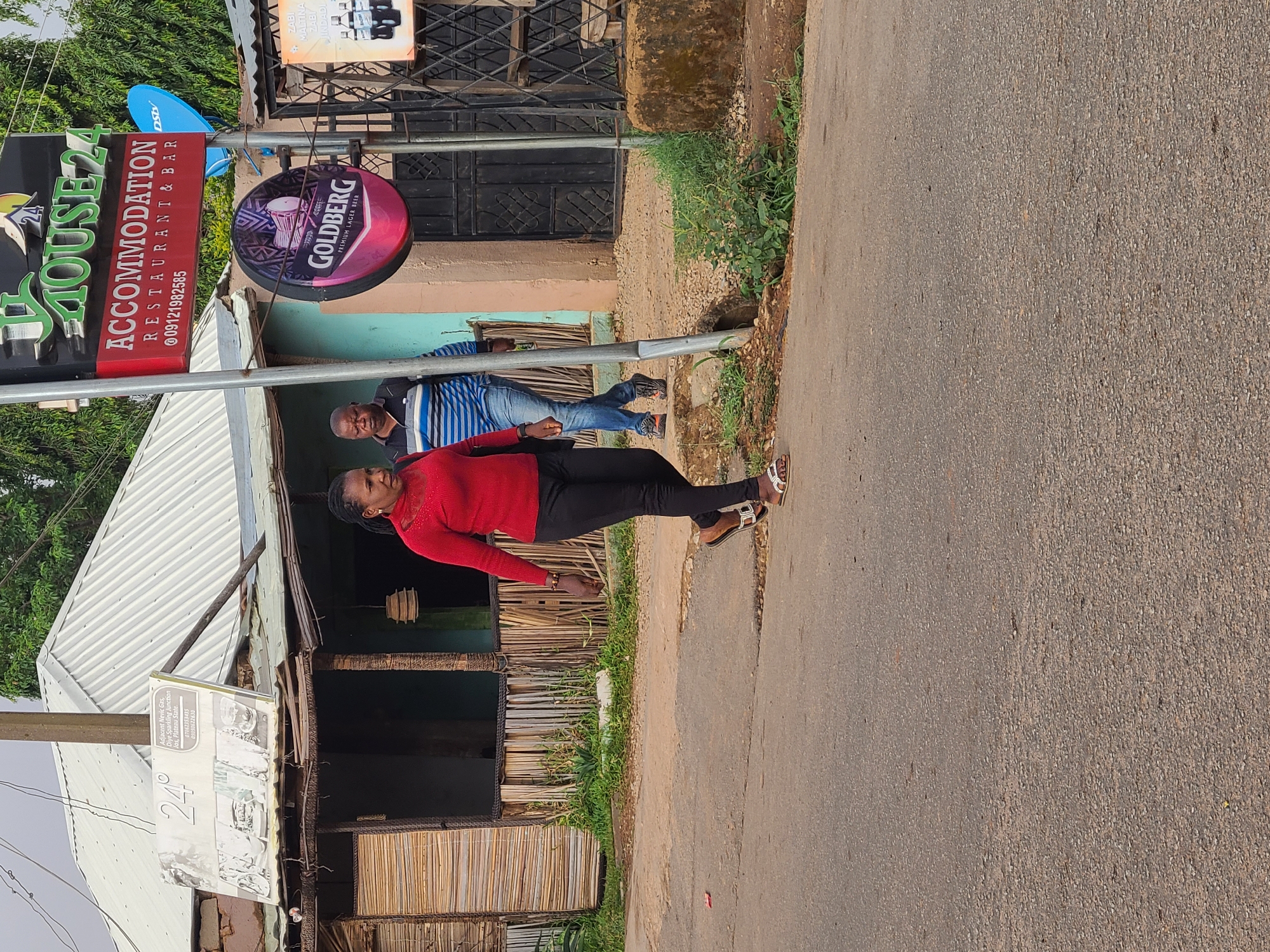 A daytime, outdoor scene in Jos, Plateau State, Nigeria, features a narrow paved road with roadside businesses. A woman in a red sweater and black pants stands on the road. Behind her, under the awning of a building, a man in a blue striped shirt is visible. The businesses include "24 House Accommodation, Restaurant & Bar," a Goldberg Premium Lager Beer advertisement, and a sign for "Adjacent Nevic Gas, Diye Sparkling Junction,". The building has a white corrugated metal roof and bamboo fencing. An antenna for DSTV is also visible.