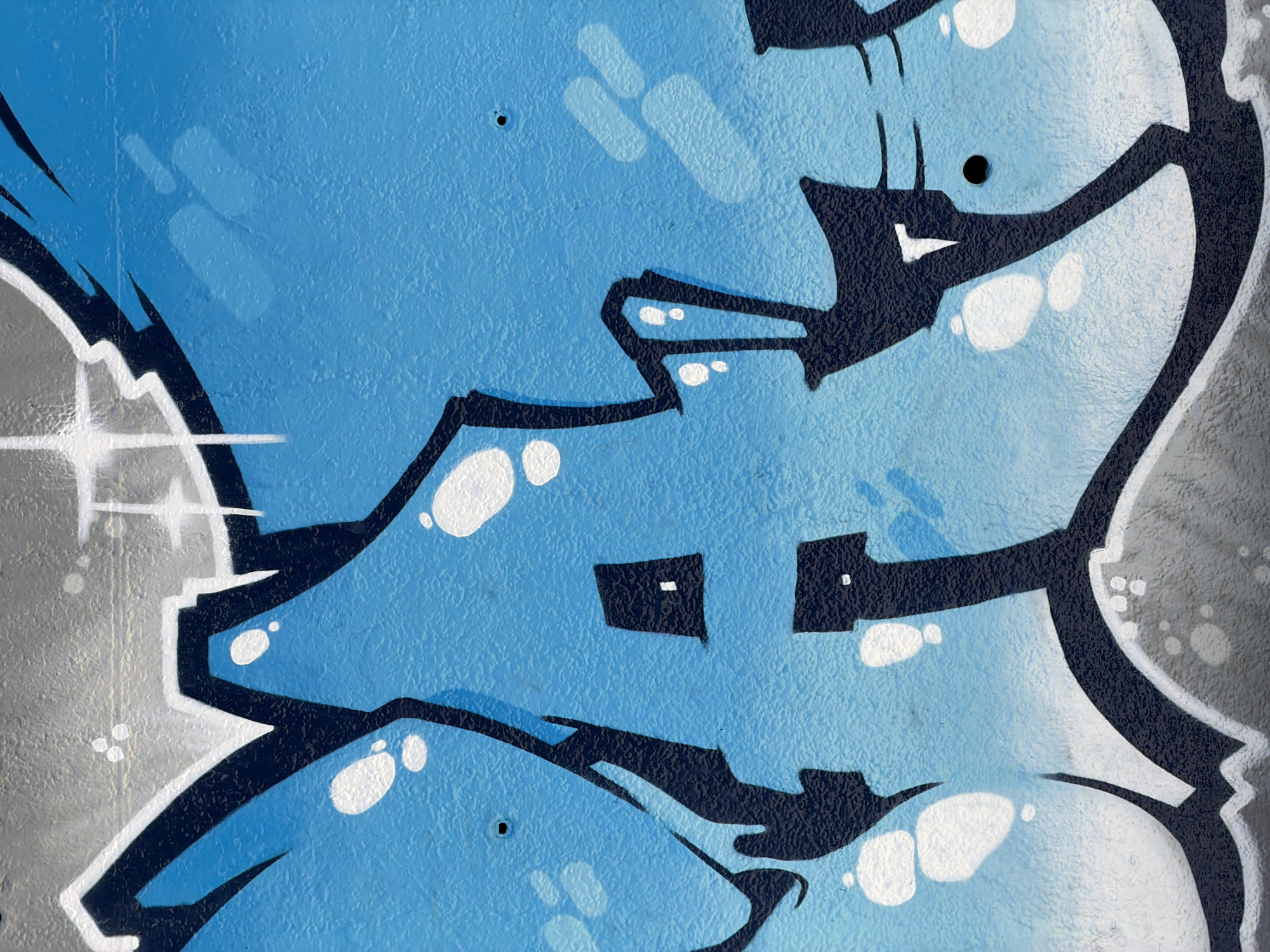 This is a close-up, vertical, eye-level, full-frame photograph of a section of graffiti art on a textured wall. The graffiti features bold, black outlines with a gradient of bright blue and white fill. Several white circular highlights are present within the blue areas, adding depth and a sense of luminescence. The grey background of the wall is visible in the upper and lower portions of the image, with some darker grey and white spray-painted accents. The graffiti appears to form abstract letter shapes. There are no people or discernible actions taking place. The image focuses solely on the artwork itself, providing no information about its specific location in Barcelona, Spain.