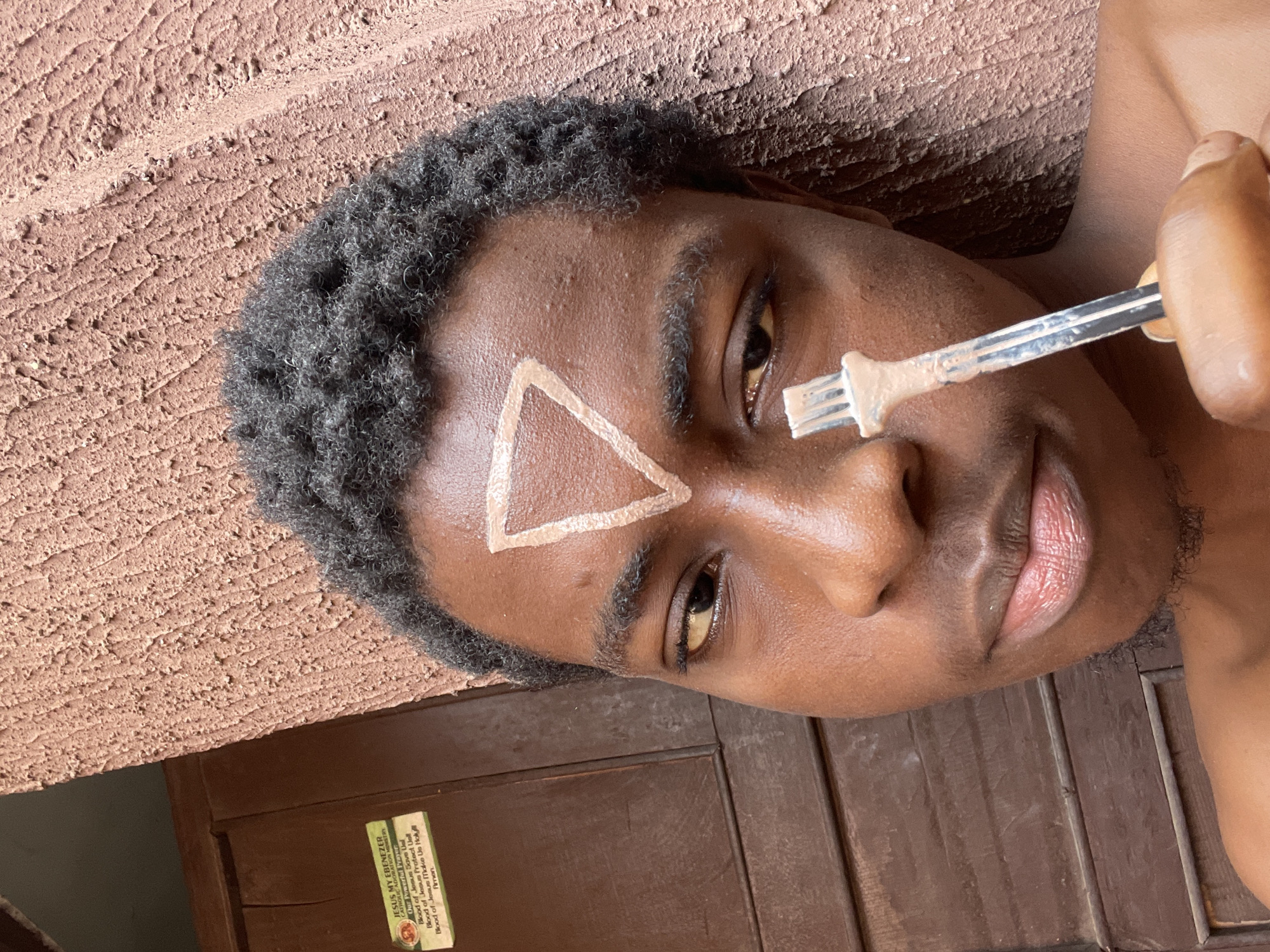 The image shows a young man with dark skin and short, curly black hair. He is positioned against a textured wall with a brown hue. A light-colored triangle is painted on his forehead. He holds a metal utensil with light brown substance on the tips, close to his face. A wooden panel is visible below the wall, bearing a sign with the text "JESUS MY EBENEZER" and other printed details. The location is implied to be Nkpor, Nigeria.