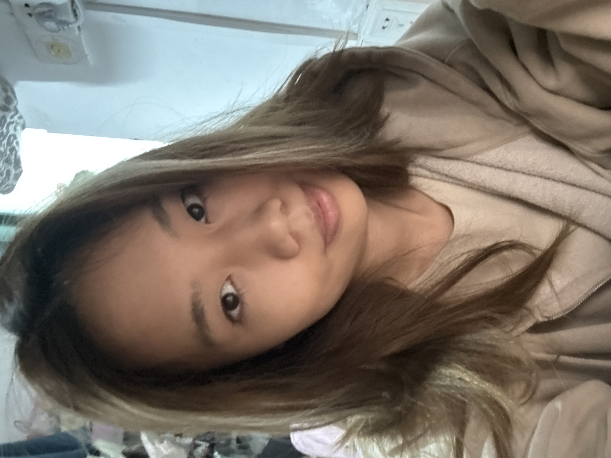 A selfie of a woman with brown hair wearing a beige hooded sweatshirt. The location is likely indoors, given the artificial light and ceiling fixture visible in the upper left corner of the frame. There is no information to pinpoint the location to Nan'an District, China.