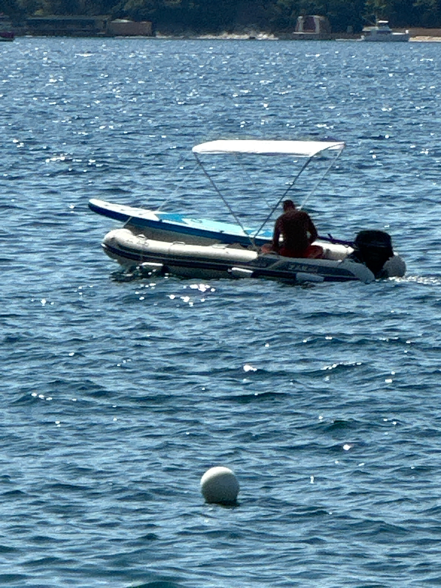An overhead view shows a small inflatable boat with a canopy moving through the blue sea. A person, silhouetted and wearing dark clothing, sits in the boat facing away from the camera, and a paddleboard is secured on top of the inflatable raft. To the right of the boat, a black outboard motor is visible, propelling the vessel forward. A white spherical buoy floats in the water in the foreground, and the distant shoreline features lush green trees and some structures, including buildings and a white boat docked on the land. The water is choppy, with sunlight reflecting off the surface, creating a shimmering effect.