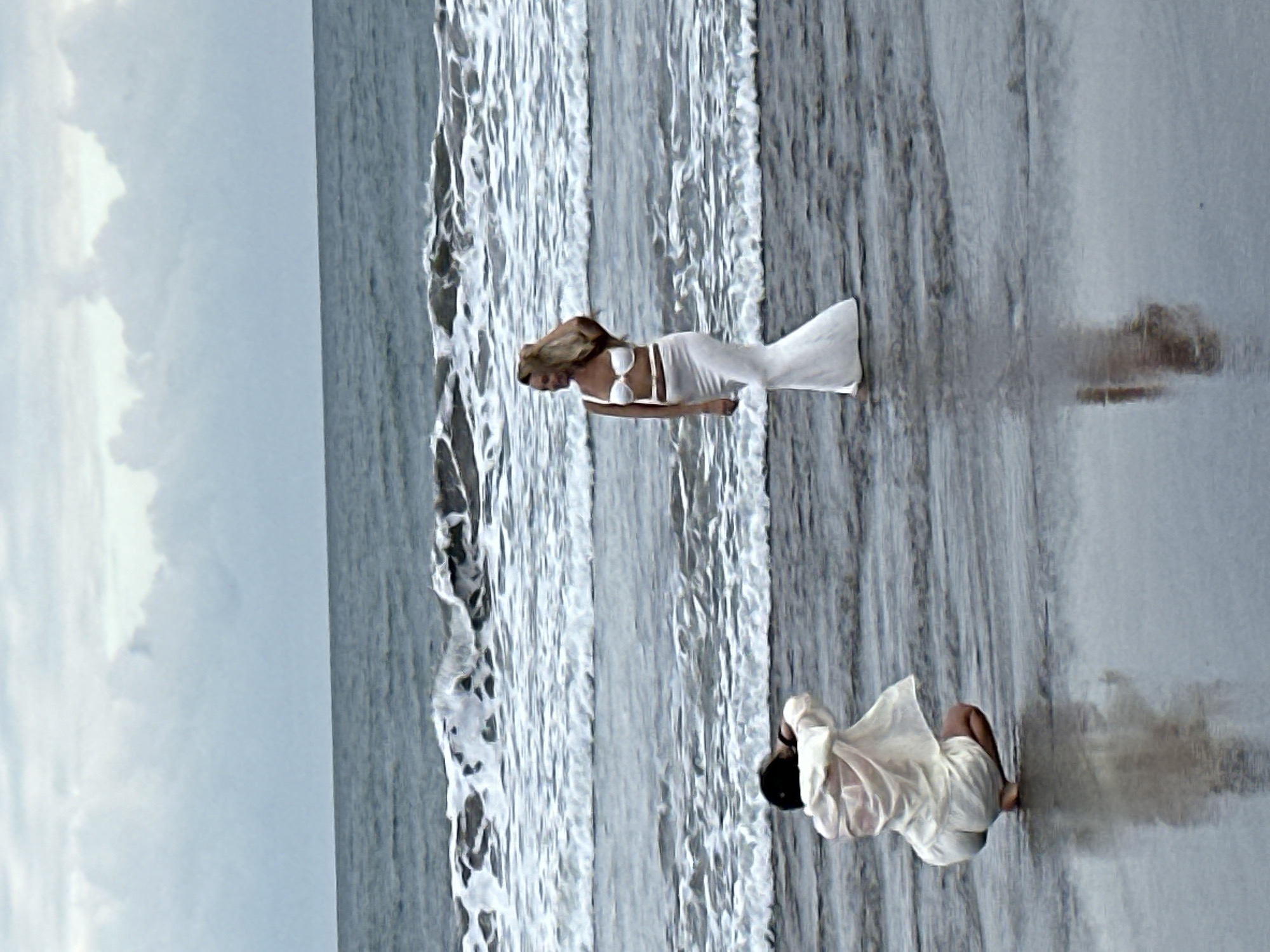 A photographer kneels on a wet beach in North Kuta, Indonesia, facing a woman posing in the ocean. The photographer, wearing a white shirt and white shorts, is crouched low and appears to be holding a camera. The woman, dressed in a white bikini top and a long white skirt, stands in shallow water, her hand raised to her hair. Waves crash behind her, and the sky is overcast. The scene is reflected in the wet sand.