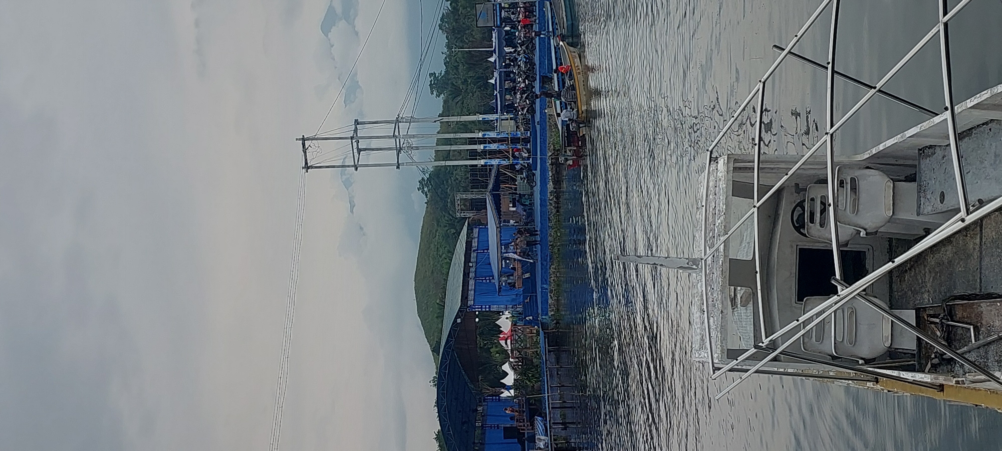 The image depicts a waterfront scene in Asei Besar, Indonesia, likely during an event. In the foreground, the bow of a white and silver aluminum boat is visible, its interior featuring two white bucket seats, a steering wheel, and a dark opening. The boat is on calm, grey water that reflects the overcast sky.
In the mid-ground, a blue stage structure with a large roof extends along the shoreline. Several white tents are set up behind the stage. A crowd of people can be seen gathered on and around the stage area. To the right of the stage, a large, multi-story metal tower with wires stretches upwards, and further to the right, a densely packed area of motorcycles is visible, suggesting a public gathering or festival.
Beyond the stage, a verdant, hilly landscape rises, with a prominent green hill topped by a small structure. In the background, misty mountains are visible through the clouds. Several smaller boats are also present on the water. One yellow boat in the mid-right of the frame has several individuals on board. The overall atmosphere appears subdued due to the cloudy weather.