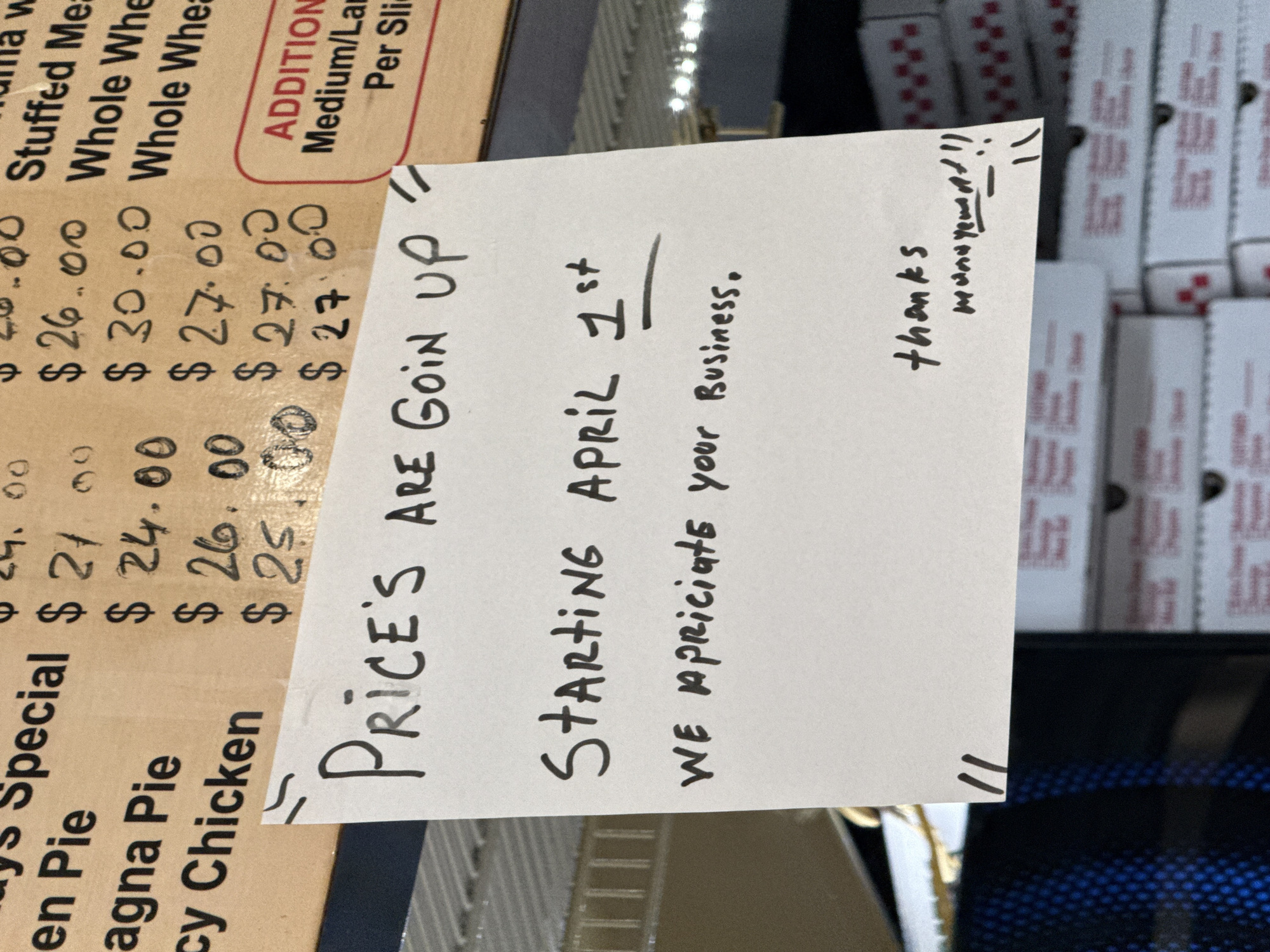 The image displays a close-up, static scene inside a food establishment located in New York, United States of America. A white sheet of paper with handwritten text in black marker is taped over a portion of a printed menu or price list. The handwritten sign reads: "PRICE'S ARE GOIN UP // STARTING APRIL 1st // WE apreciate your Business. // thanks // management!!" Two double-slash marks are drawn above and below the main message on the white sign.
Partially visible behind and above the sign is a light-brown printed menu. On the left, item names like "en Pie", "Lasagna Pie", and "cy Chicken" are discernible. On the right, corresponding prices ranging from $21.00 to $30.00 are listed, some appearing to be handwritten modifications or updates. Text on the far right of the menu includes "ADDITIONAL" and "Medium/Lar Per Slice". Below the sign, stacks of white cardboard boxes with red branding, identifiable as "FIVE GUYS" packaging, are visible. To the far left, a blue perforated surface and a metallic rack are partially visible, suggesting a refrigeration unit or shelving. No individuals are present in the scene.