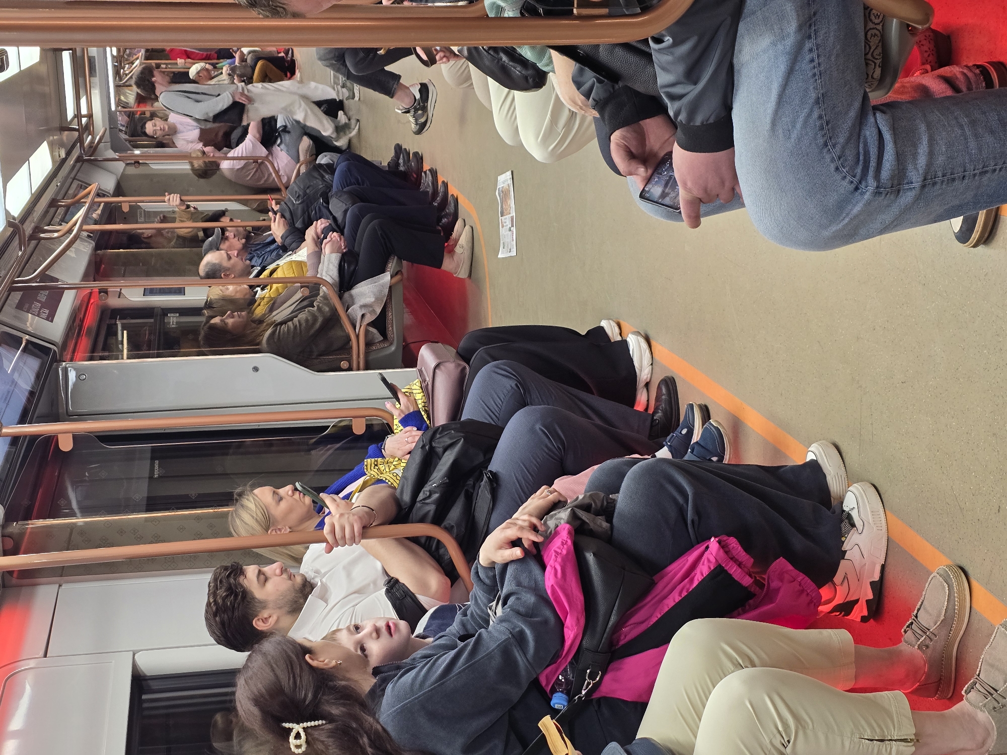 The image captures a scene inside a train or subway car. Passengers are seated on either side of the aisle, some holding onto the handrails. People are seen using their phones. A newspaper lies on the floor of the train car. The location is not specified.
