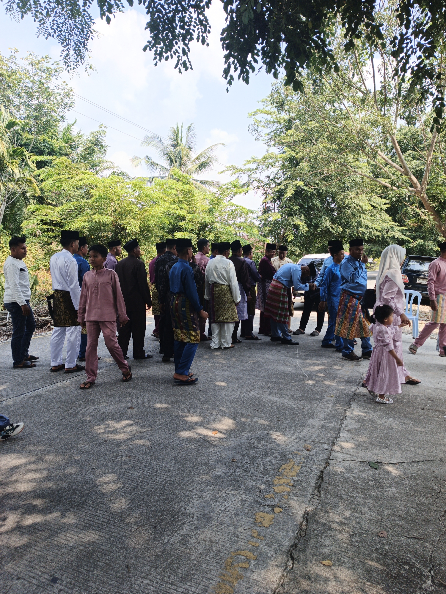 In Pematang Duku, Indonesia, a group of men and a young girl are gathered on a paved area, possibly a street or pathway, with trees and foliage in the background. The men are mostly dressed in traditional attire, including peci hats and sarongs. Some are standing and looking in various directions, while others appear to be interacting or conversing. One man in the foreground is wearing a light pink traditional outfit. A young girl in a similar pink dress is walking to the right side of the frame. The environment suggests an outdoor setting with dappled sunlight filtering through the trees, creating shadows on the ground. Two cars are partially visible in the background.