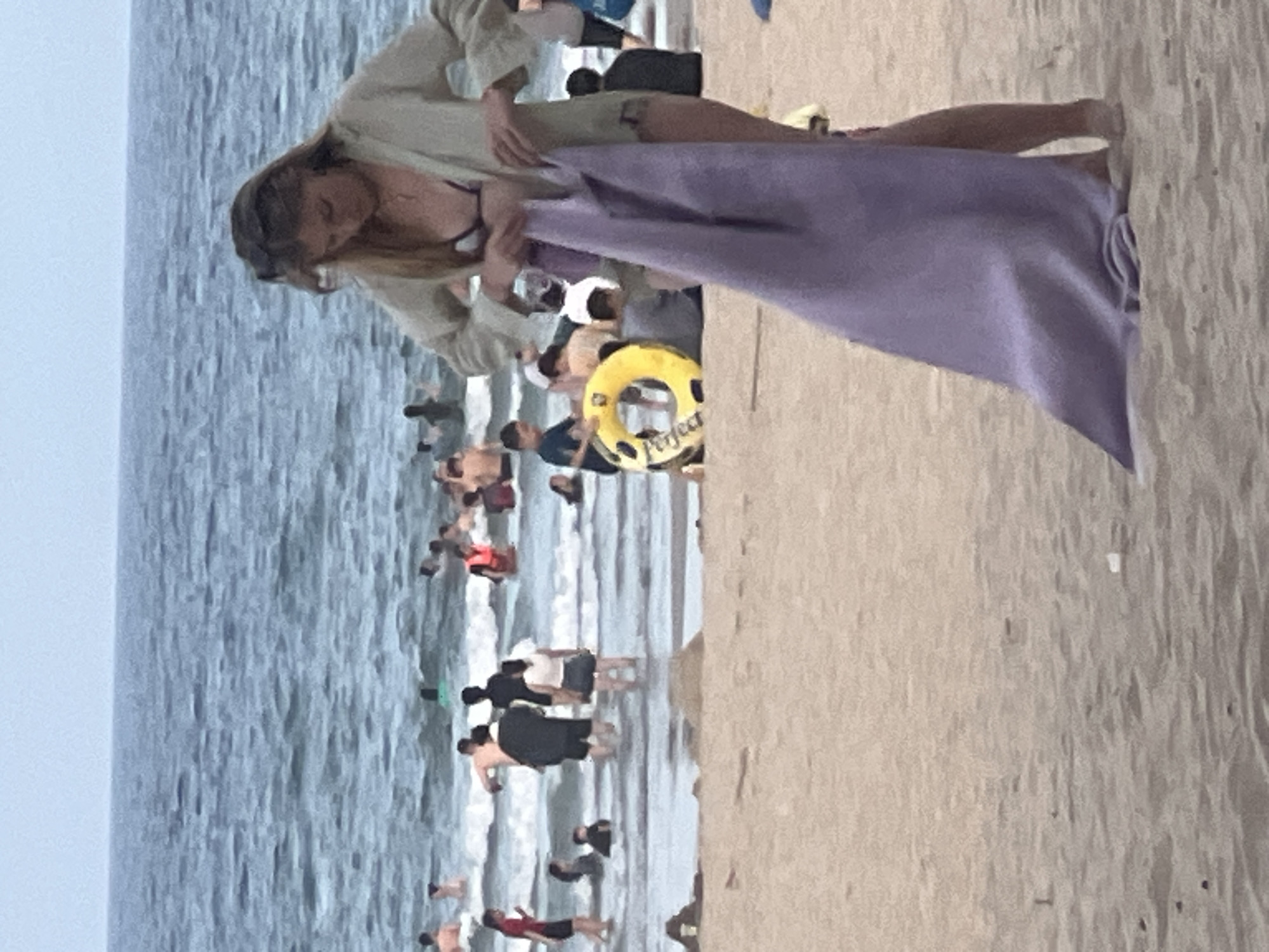 The media file depicts a crowded beach scene at Đà Nẵng, Vietnam. The primary subjects are numerous people enjoying the ocean and the sandy shore. A woman in the foreground, wearing a light-colored top and a bikini top, is seen holding a large purple towel. She appears to be preparing to dry off or cover herself.
In the water, many individuals are wading, swimming, and interacting. Some are partially submerged in the waves, while others are standing closer to the shore. Among the beachgoers, a yellow inflatable ring with the word "Perfect" printed on it is visible, suggesting recreational activities. The overall atmosphere appears lively, with a sense of summer leisure and social gathering.
The scene is set on a beach with calm, greyish-blue water and a sandy shoreline. The sky is overcast, indicating likely cloudy weather. The presence of numerous people in swimwear and engaging in beach activities points to daytime, possibly late afternoon. The location cues, such as the distinct beach environment and the mention of Đà Nẵng, Vietnam, confirm the setting. While the image is somewhat pixelated, it captures the essence of a busy beach day. There are no overt signs of specific emotions, but the general ambiance suggests a relaxed and enjoyable experience for the beachgoers. The visible text "Perfect" on the inflatable ring is the only discernible text in the image.