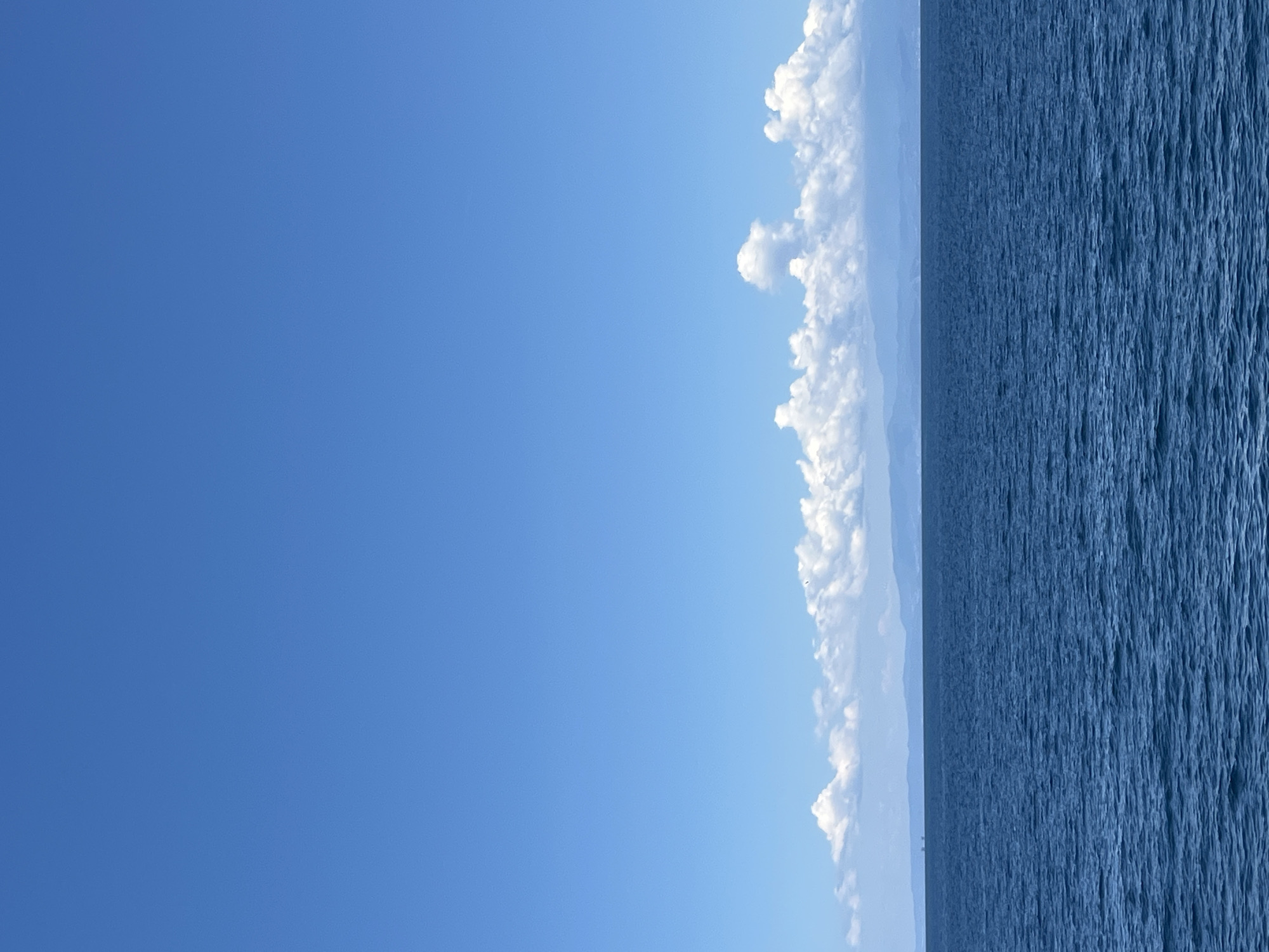 An expansive, clear blue sky dominates the upper half of the image, with the horizon line appearing approximately one-third of the way up from the bottom. Below the sky, a band of white, puffy cumulus clouds stretches across the width of the frame. These clouds sit above a distant, hazy landmass that is likely the coastline or mountains of Thassos, Greece. The lower half of the image is filled with the deep blue, slightly rippled surface of the sea. There are no people or prominent objects visible in the water or on the land. The scene conveys a sense of calm and vastness.