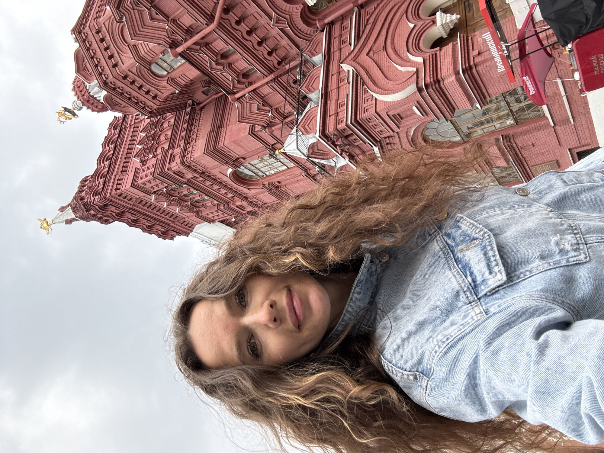 A woman with long, curly brown hair takes a selfie in front of a large, ornate red brick building with golden spires. The building appears to be a historical landmark, possibly a museum or government building, with intricate architectural details. The woman is wearing a light blue denim jacket. The sky is overcast, and the scene suggests an outdoor setting in a city. The text visible on a red canopy in the background is in Russian, and appears to be related to a vendor or stall. The building's style is consistent with architecture found in Moscow, Russia, particularly around Red Square.