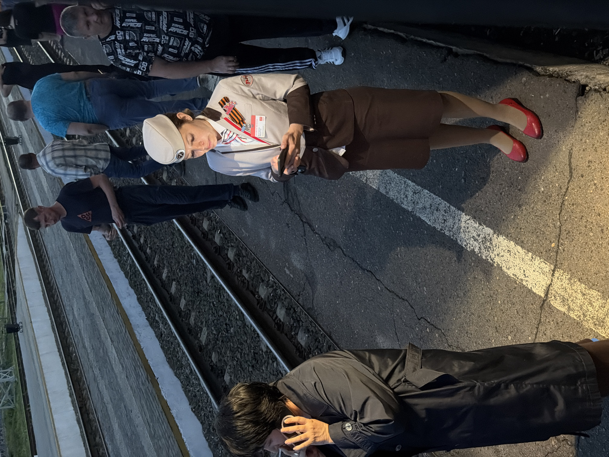 The image shows a group of people standing near railroad tracks. They are standing on an asphalt surface with painted white lines. The group includes a woman in a light gray uniform jacket, brown skirt, and red shoes, wearing a white peaked cap and the St. George ribbon. The woman is holding a small black object. Other people in the group are wearing casual clothing. The location is likely in Rtishchevo, Russia, given the person wearing the St. George ribbon.