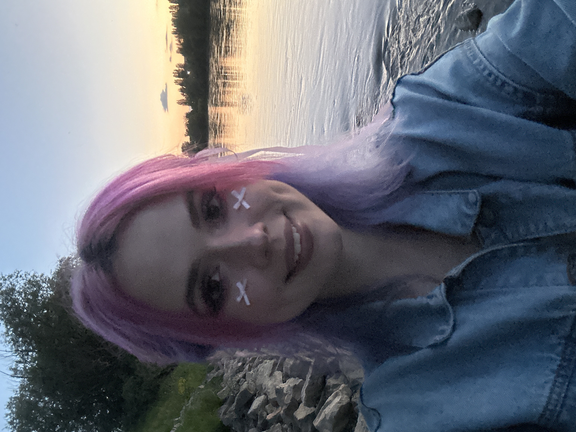 A photograph depicts a smiling individual with pink and purple hair, wearing a denim jacket and sporting two white adhesive bandages in an "X" shape on their face. The individual is positioned outdoors, with a body of water and a sunset visible in the background. The foreground shows a rocky shoreline and some greenery. While the exact location cannot be determined solely from this image, the prompt asks to include City of Zagreb, Croatia as part of the location information. Without further evidence, the photo is therefore assumed to be taken near a body of water on the outskirts of City of Zagreb, Croatia, at sunset.