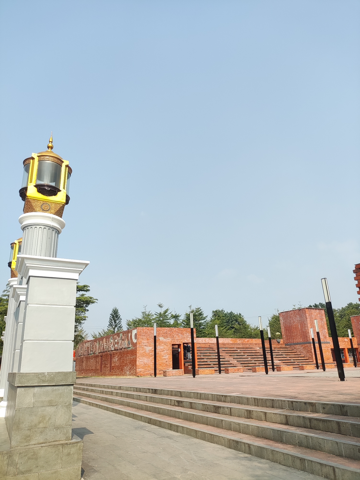 This is a low-angle, outdoor, full shot of a public space in Majalengka, Indonesia. In the foreground, to the left, is a tall, white, and grey column with a decorative, golden lantern on top. To the right of the column are paved stairs leading up to a large, open, brick-paved area. In the background, there are brick buildings, tiered seating areas, and several tall, black, pole-like lights. A clear, light blue sky fills the upper portion of the image. The scene appears to be empty of people.