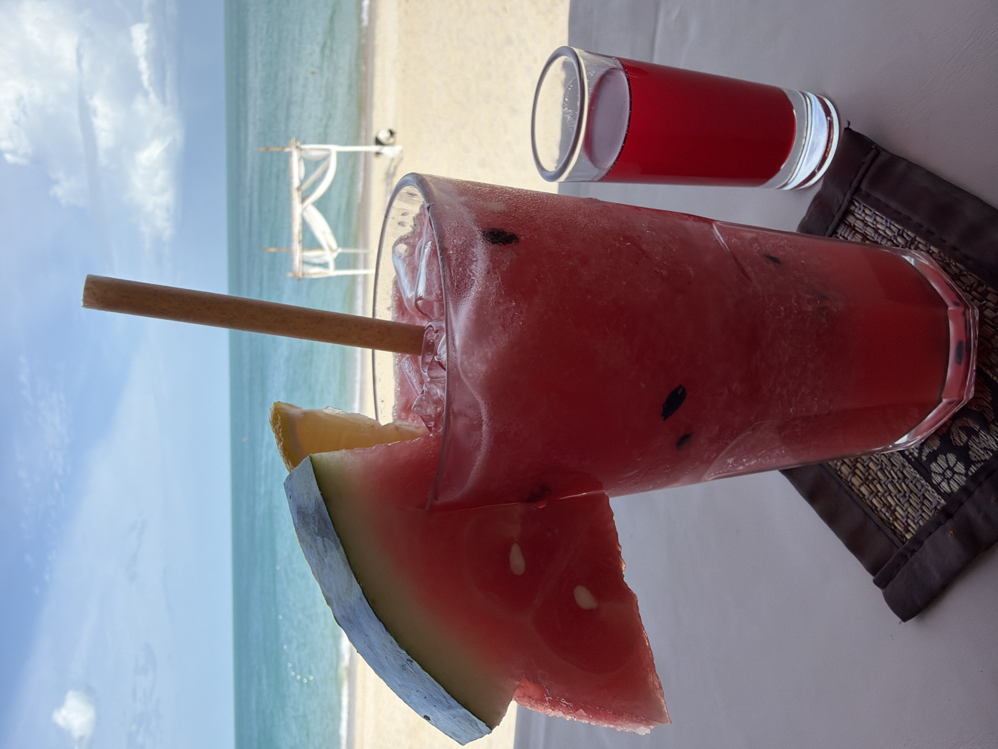A tall glass of red juice, likely watermelon, is garnished with a slice of watermelon and a yellow wedge, possibly lemon, and a bamboo straw. Next to it, a small shot glass contains a darker red liquid. Both glasses are on a table with a decorative dark placemat. In the background, a beach and the ocean are visible under a partly cloudy sky, with a white gazebo structure on the sand. The scene suggests a relaxed setting in the coastal area of Ko Samui District, Thailand.