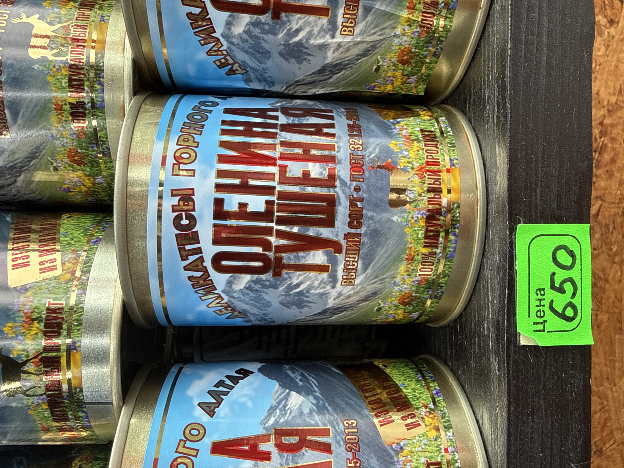 A close-up, eye-level, outdoor shot features a shelf displaying several cans of canned meat. The cans are cylindrical with colorful labels depicting mountainous landscapes and grazing animals. The text on the labels is in Russian. One can is prominently labeled "ОЛЕНИНА ТУШЕНАЯ" (Stewed Venison). A bright green price tag with the word "Цена" (Price) and the number "650" is affixed to the shelf in the lower-left corner. The shelf appears to be made of dark wood. The background is blurred, suggesting a market or store environment. The scene is likely located in the city of Manzherok, Russia, given the context of the canned goods and their labels.