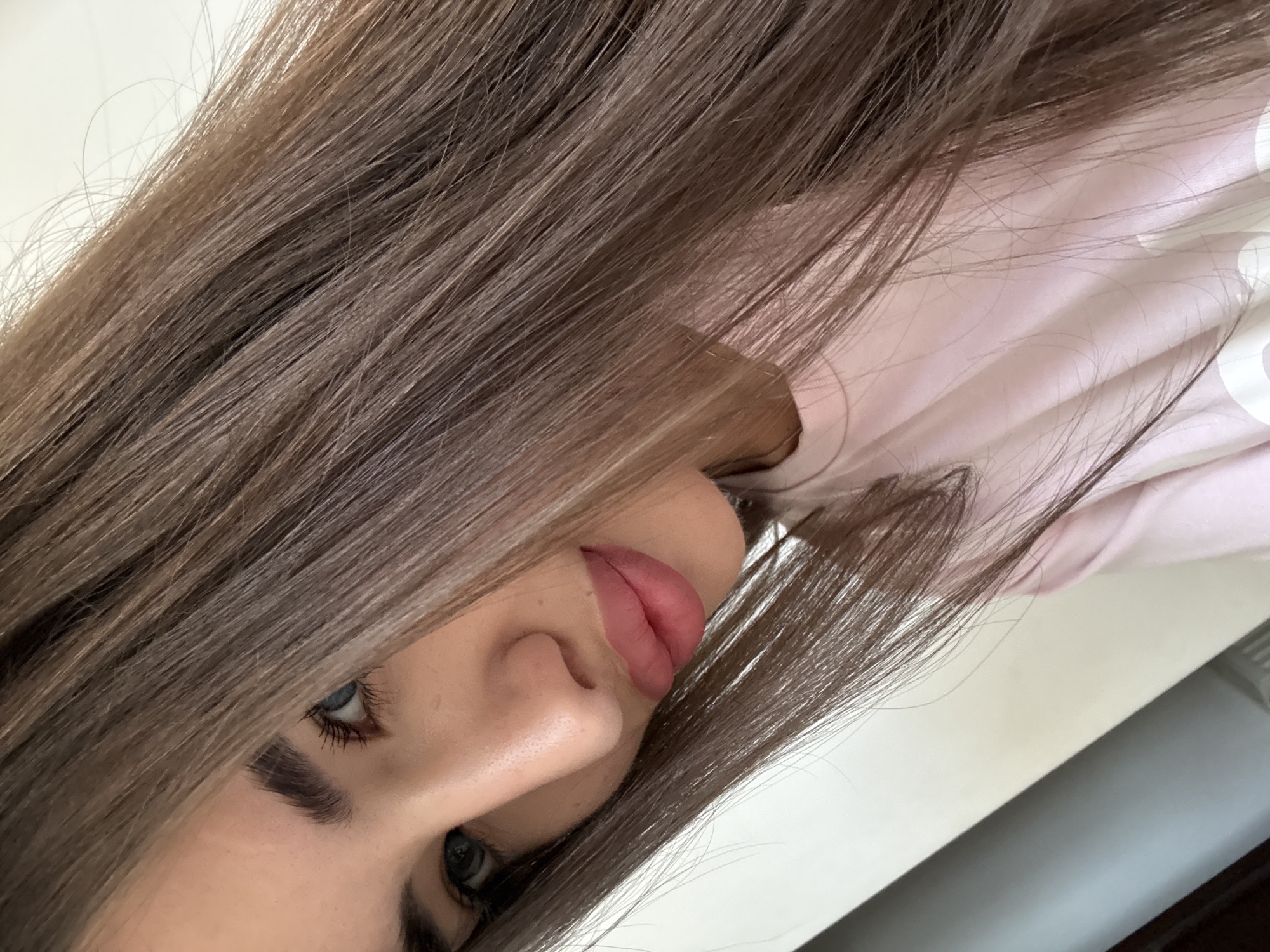 A close-up selfie of a young woman with long, straight brown hair framing her face. She has striking blue-green eyes, defined eyebrows, and full lips with pink lipstick. She is wearing a light pink t-shirt. The background is a plain, off-white wall with a diagonal shadow. The lighting suggests an indoor setting. No specific location in Chișinău, Moldova can be identified from the image alone.