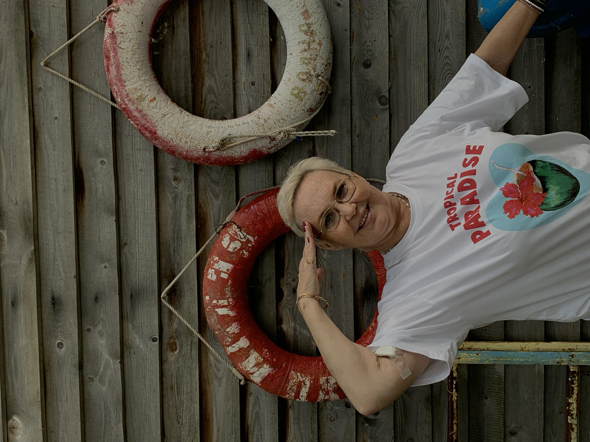 In Chaikovsky, Russia, a fair-skinned woman with blonde hair and glasses, wearing a white t-shirt with a "Tropical Paradise" graphic, salutes while standing in front of a weathered wooden wall. Two red and white life preservers hang on the wall behind her. The woman has a gold bracelet and a ring on her right hand, and a bandage is visible on her upper left arm. The overall scene appears to be outdoors, possibly in a recreational area or near a body of water.