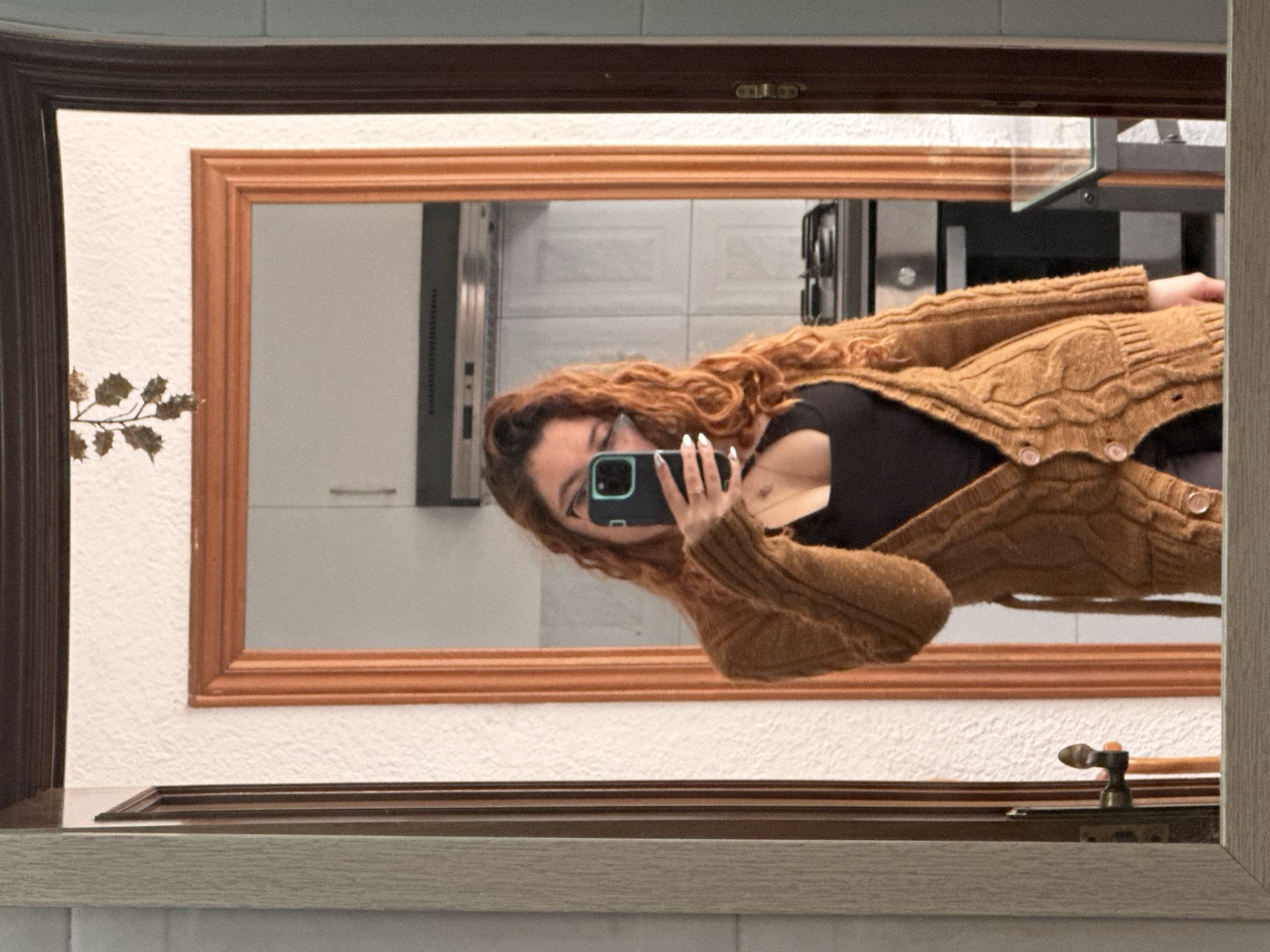 A woman is taking a selfie in a mirror. She has curly red hair and is wearing a black top and a brown cardigan. The mirror is framed in brown and wood, and reflects a tiled wall and kitchen appliances. The photo appears to be taken in a bathroom or kitchen, but it is difficult to know if it is in the city of Barcelona, Spain based on the available details.