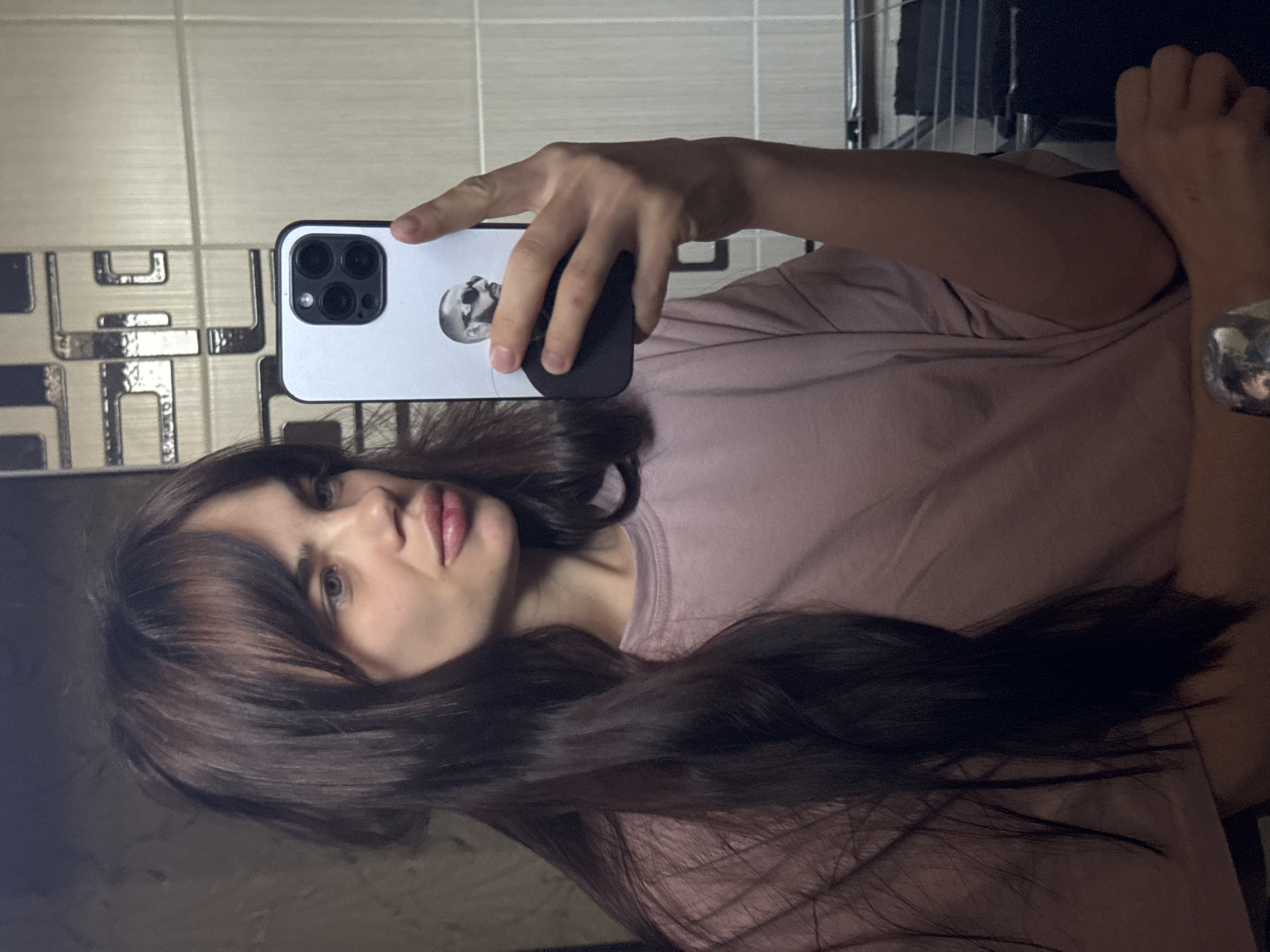 A woman takes a selfie in a bathroom with a modern tiled wall. She is holding a smartphone with a white case featuring a design of a man's face with sunglasses. The woman is wearing a pink t-shirt, and her dark brown hair is styled with bangs. The location is likely inside a private residence. There is no information suggesting the image was taken in Rostov-on-Don, Russia.