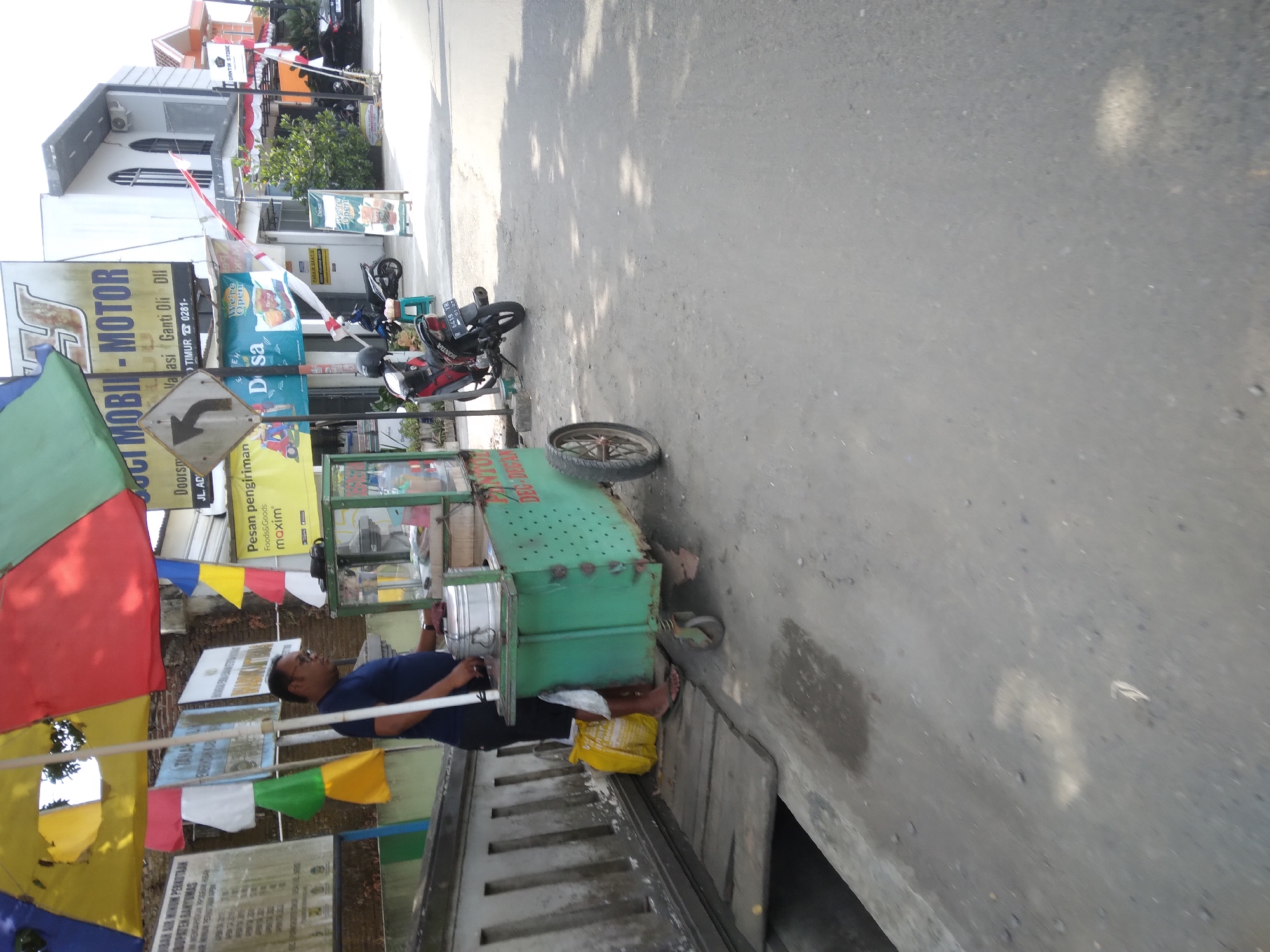 An outdoor, eye-level, medium shot captures a street vendor and his food cart in Sokaraja, Indonesia. The vendor, a man wearing a dark blue t-shirt and dark shorts, stands by his green cart, which is laden with food. He is looking down, seemingly engaged in preparing or serving food. The cart has a canopy of colorful umbrellas overhead, featuring shades of yellow, red, and green. Behind the vendor and cart, a white building with arched windows is visible, suggesting an urban or semi-urban setting. Several parked motorcycles are also present in the background, along with various signs and flags, indicating commercial activity. The ground is paved with asphalt and dirt, with shadows cast by the sun. To the left of the cart, a yellow bag and white plastic bags are placed on a wooden platform that extends over a dark opening. A traffic sign with a left-pointing arrow is mounted on a pole near the cart. The scene appears to be set on a sunny day.