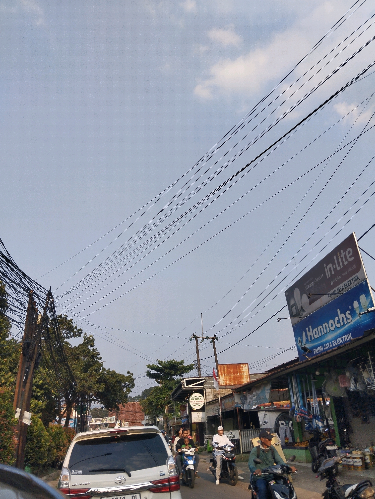 This is a photograph of a street scene in Sukaraja, Indonesia, featuring a traffic jam with several motorcycles and a car. The sky is a pale blue with scattered clouds and is crisscrossed by numerous power lines. On the right side of the frame, there are storefronts with various signs, including "in-lite" and "Hannochs" for Fahmi Jaya Elektrik. People are riding motorcycles, and a white Toyota Avanza is visible in the foreground, stopped in traffic. Trees and utility poles line the street, contributing to a sense of urban clutter. The overall impression is of a busy, everyday scene in an Indonesian town.