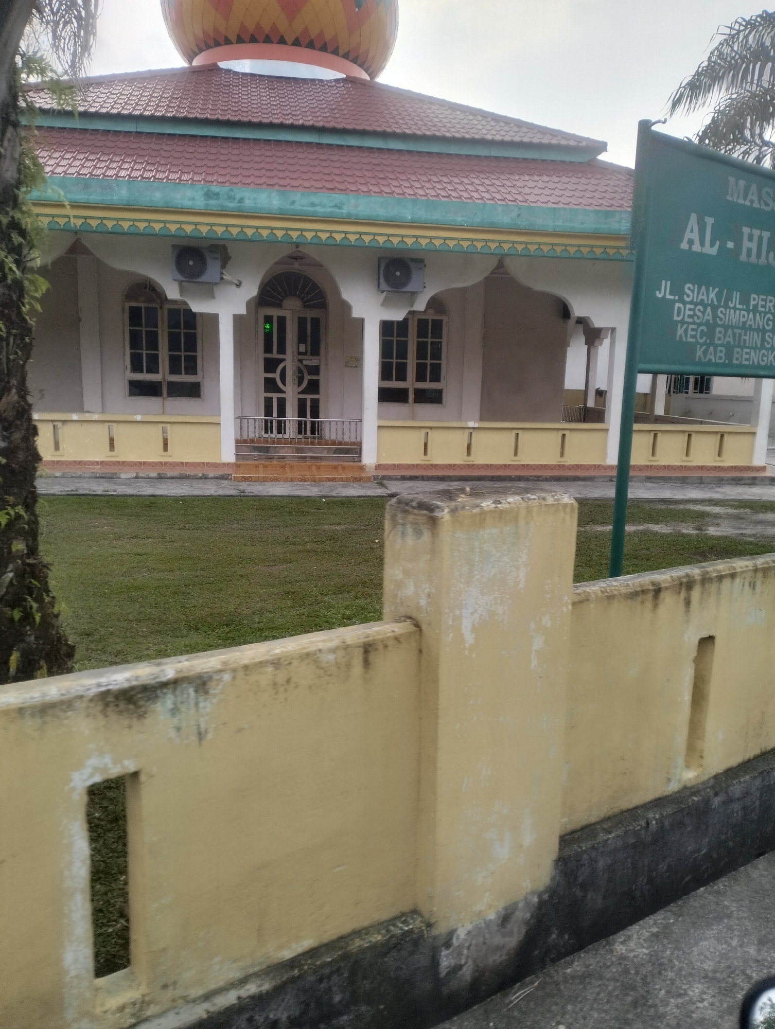 The image depicts the exterior of a mosque named AL-HIJRAH, located at JL. SIAK/JL. PERUMNAS, DESA SIMPANG EMPAT, KEC. BATHIN SOLOPAN, KAB. BENGKALIS, in Tambusai Batang Dui, Indonesia. The mosque has a red tiled roof, white walls with decorative yellow and teal trim, and large arched windows and doors. An air conditioning unit is visible on the exterior wall. In the foreground, a weathered yellow concrete wall with rectangular openings runs across the frame. A green signpost with white text indicating the mosque's name and address stands to the right of the building. The scene is set against a backdrop of green grass and trees, with a cloudy sky overhead. No people are visible in the image.