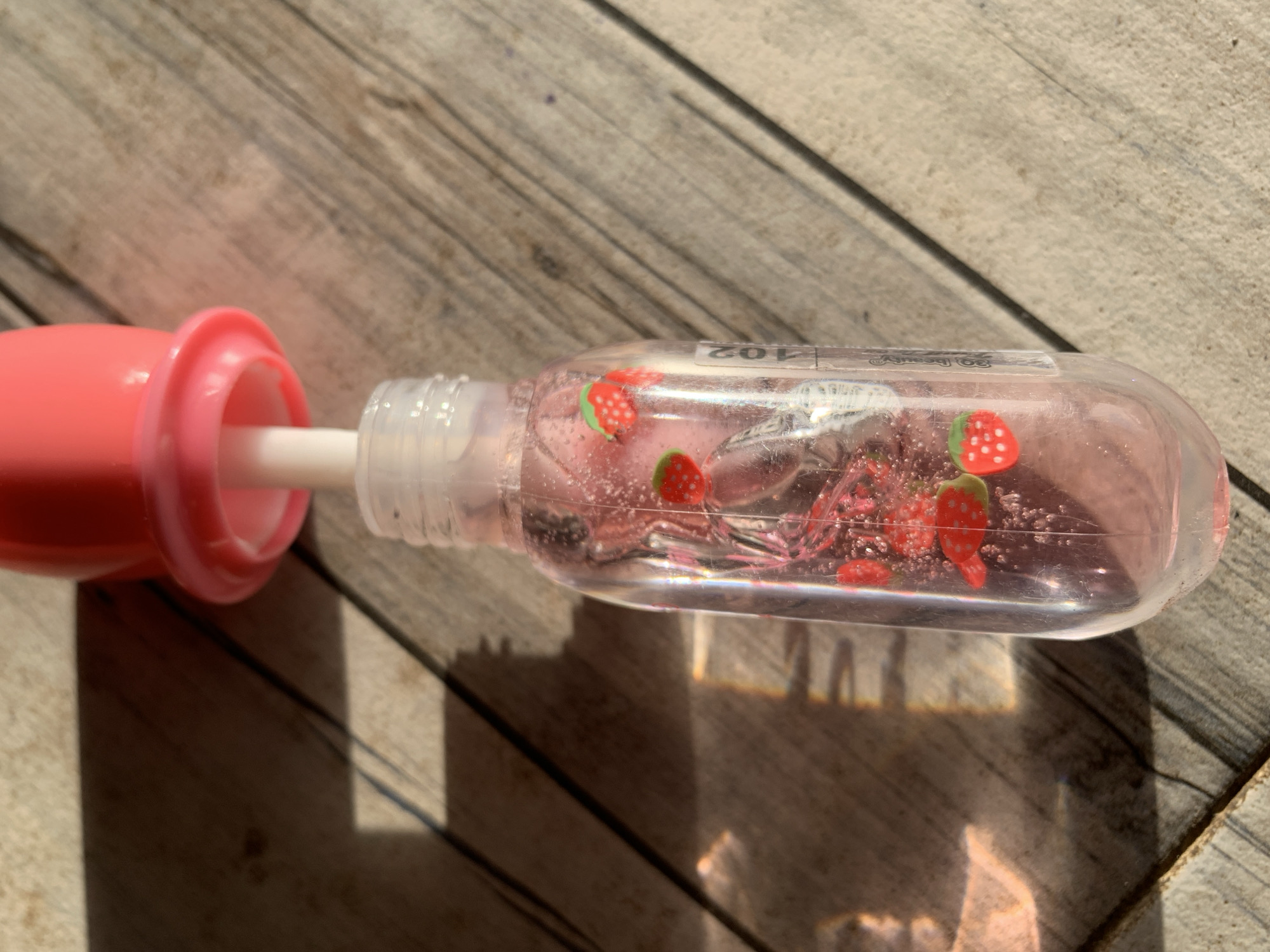 A close-up photograph displays a clear plastic container of strawberry-scented lip gloss with a pink applicator lid, resting on a weathered wooden surface. Inside the gloss are small, artificial strawberry decorations. The lighting is bright, casting shadows from the gloss container and the wooden planks. No people are visible. The setting seems to be outdoors, given the natural light. There is no discernible evidence to suggest a specific location in New Stadium, Nigeria.