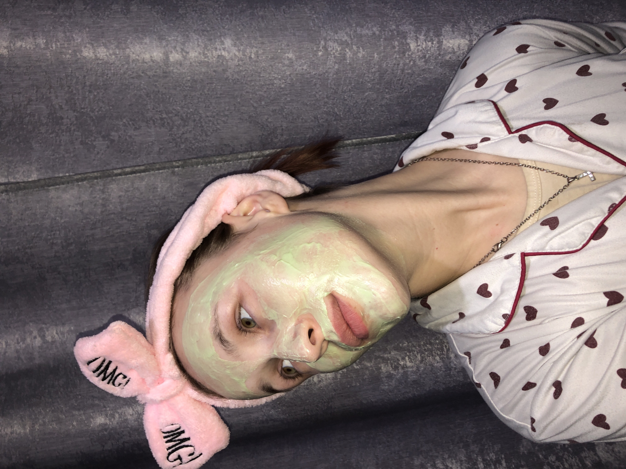 The image shows a woman lying on a grey surface. She is wearing a pink headband with "OMG!" written on it, and her face is covered in a green mask. She is also wearing pajamas with a heart pattern. A necklace is visible around her neck. The background is a textured, grey surface. There is no evidence in the image to indicate a location in the city of Фоки, Russia.